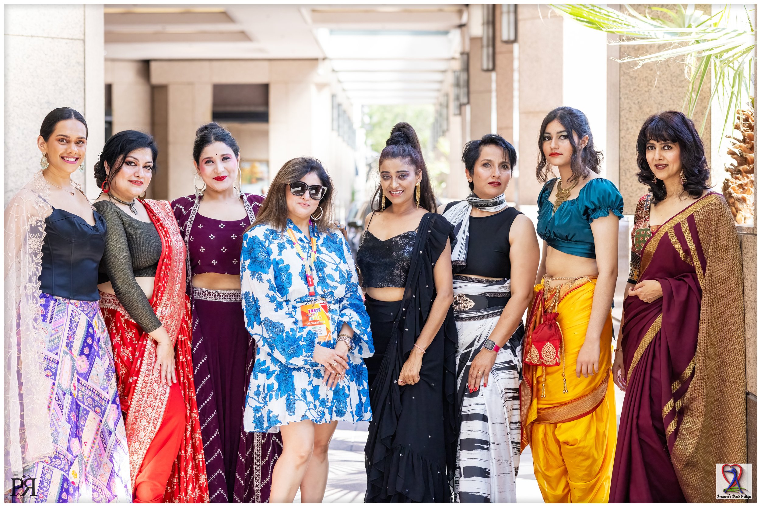 Nine women standing in a row outside, wearing colorful traditional Indian clothing and accessories, smiling at the camera.