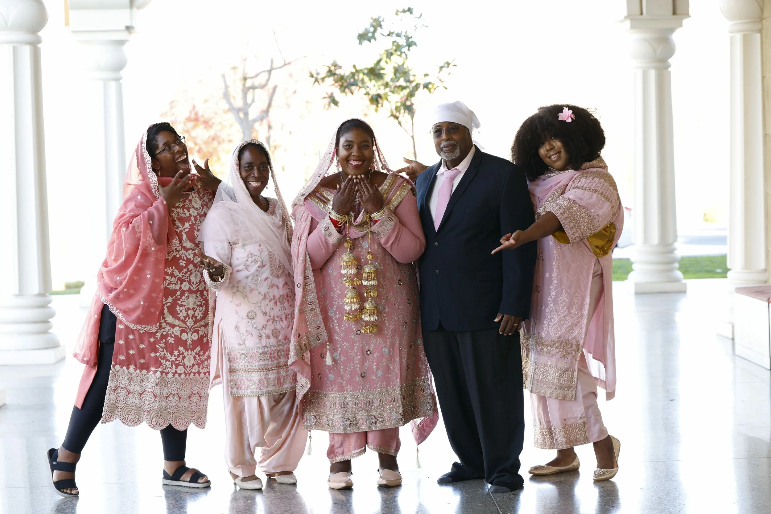 Group of six people in traditional Indian attire, standing on a porch with white columns, celebrating, smiling and posing for the photo.