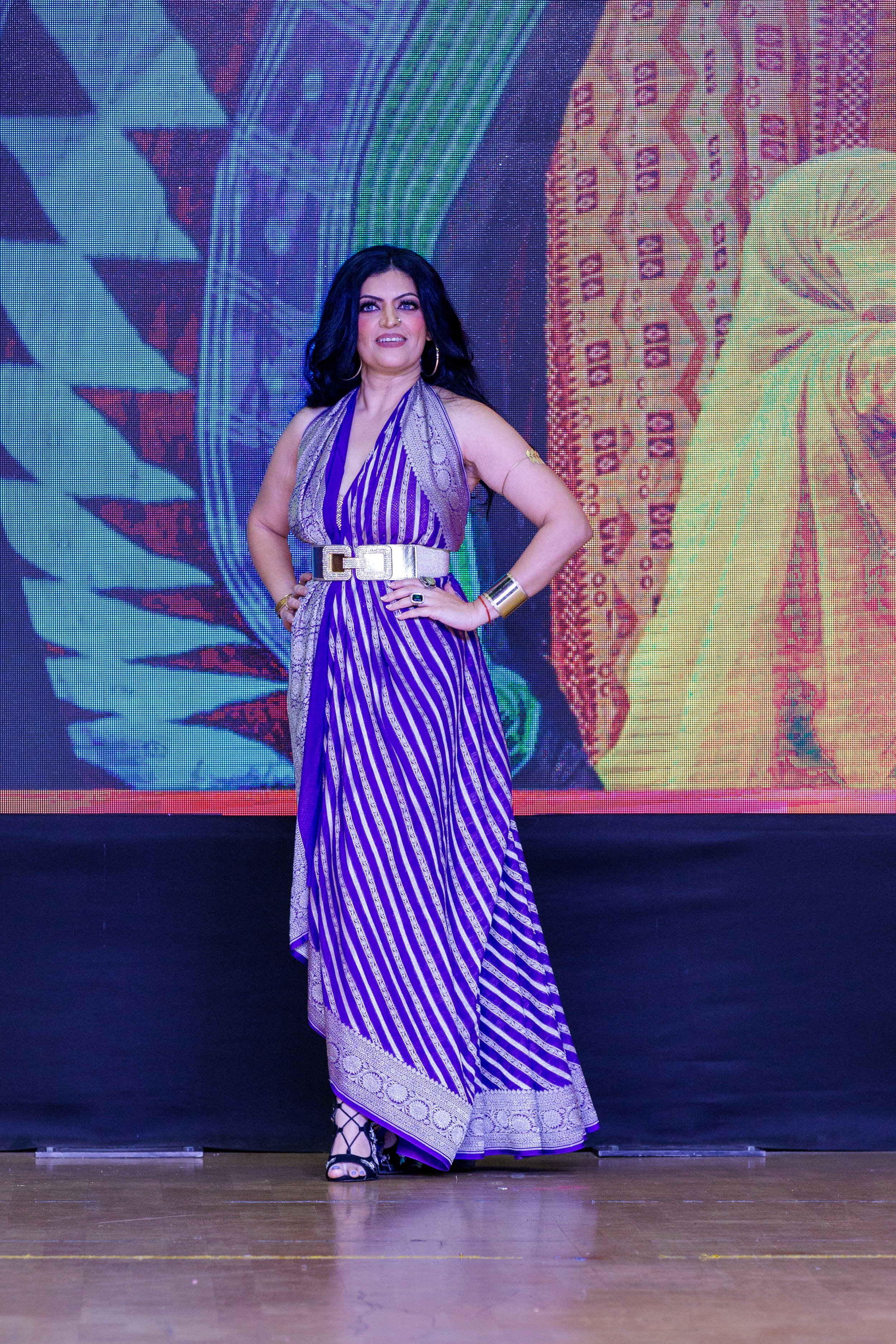 Woman with long black hair wearing a purple and white striped dress and black heels, standing on a stage with a colorful digital background.