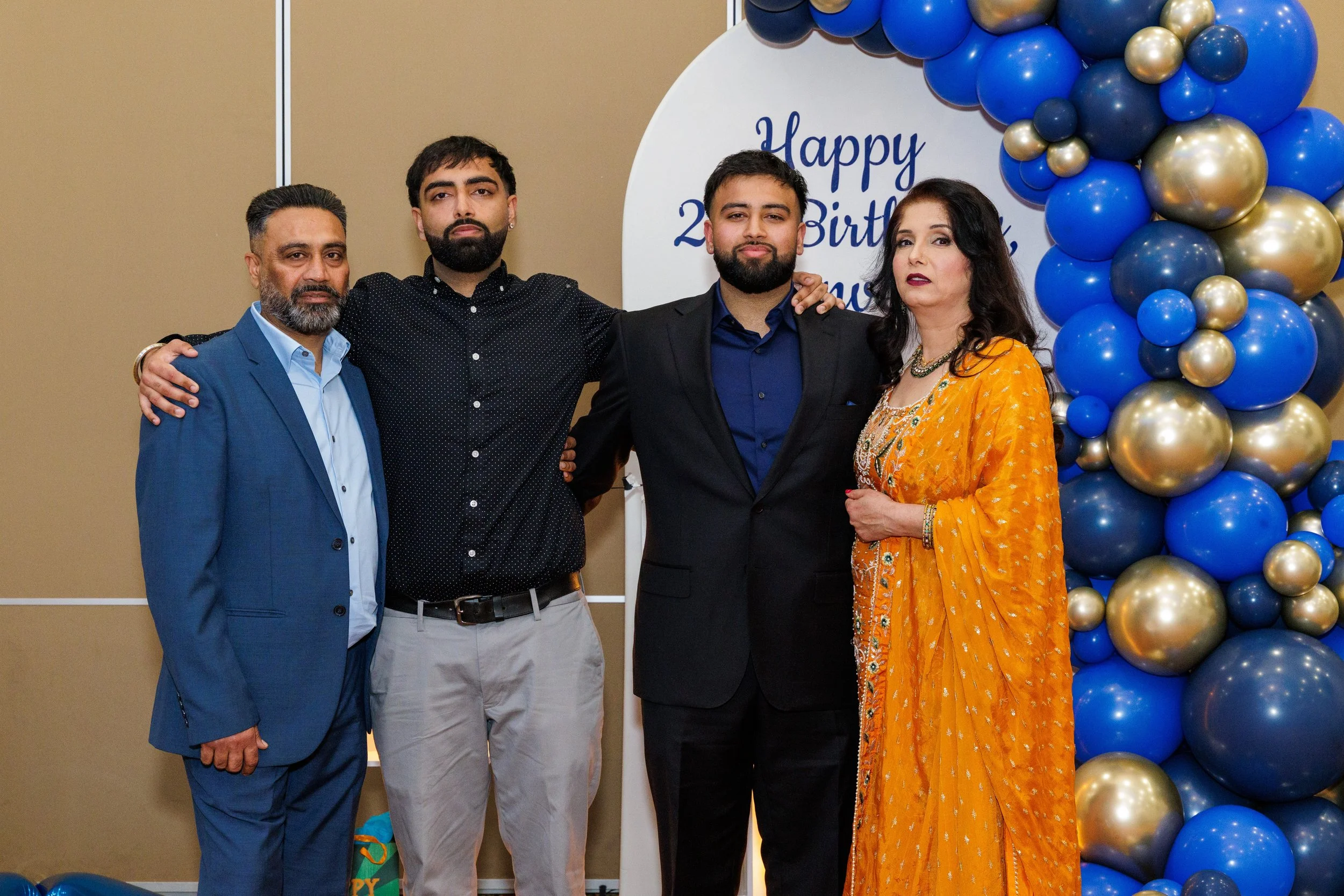 A family of five standing together at a birthday celebration, with a decorated balloon arch in blue, gold, and dark blue and a white sign with blue writing in the background.