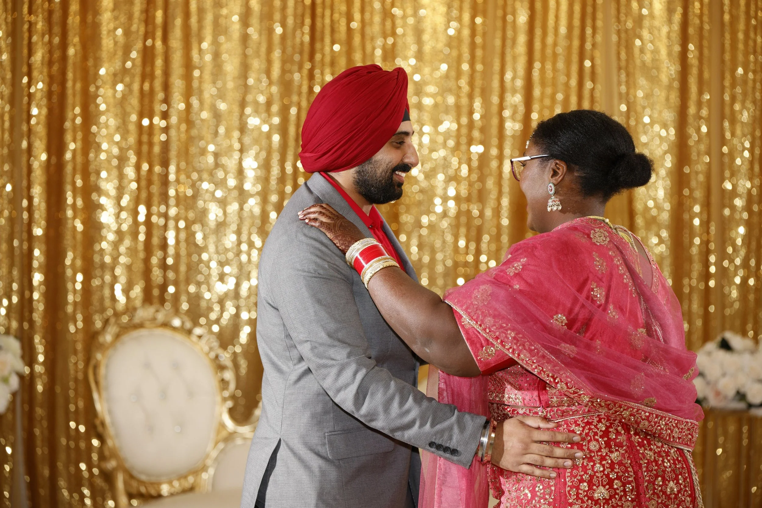 A man in a gray suit and red turban dancing with a woman in traditional Indian attire with jewelry, in front of a golden sparkly backdrop.