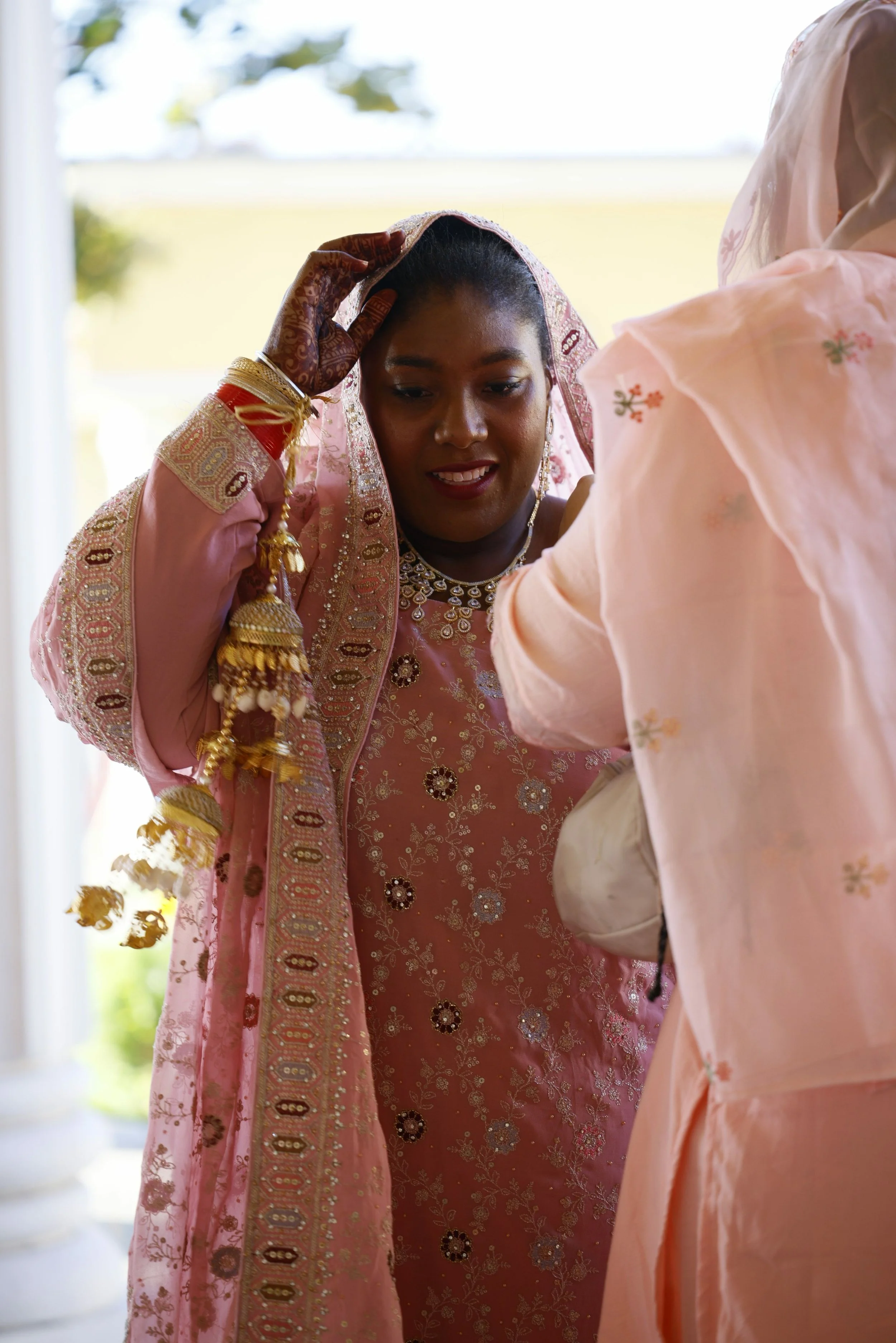 A woman in traditional Indian or South Asian attire, wearing intricate jewelry and a pink embroidered outfit, is adjusting her headscarf while another person helps her. The woman has henna on her hands and is indoors with natural light coming through