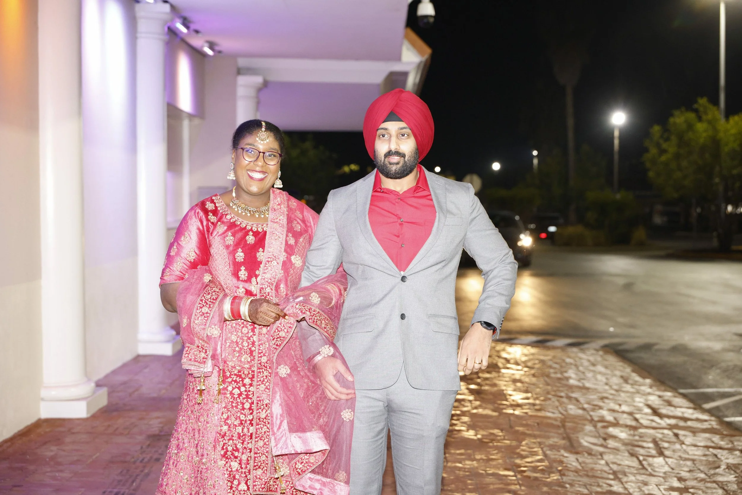 A man in a gray suit and red turban standing next to a woman in traditional pink Indian attire with jewelry outside at night.