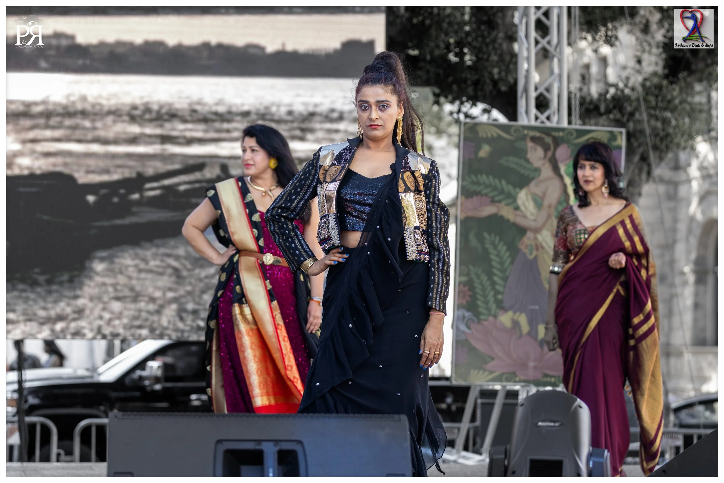 Three women in colorful traditional Indian sarees and western outfits walking on a stage during a cultural event with a large scenic backdrop and a painted canvas of a woman in traditional attire.