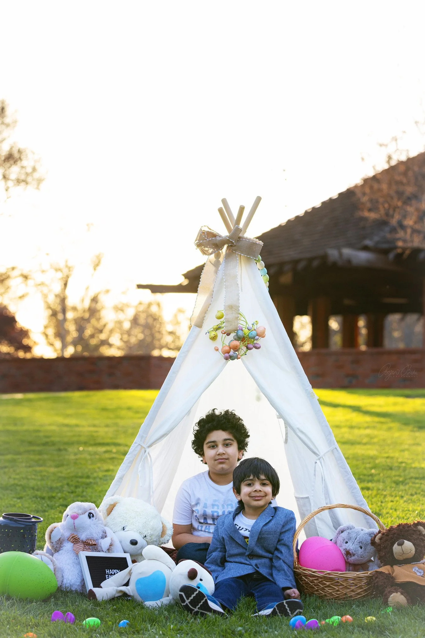 Two boys sitting in front of a small white tent decorated with pastel-colored ornaments, surrounded by stuffed animals and colorful Easter eggs, in a grassy outdoor setting during sunset.