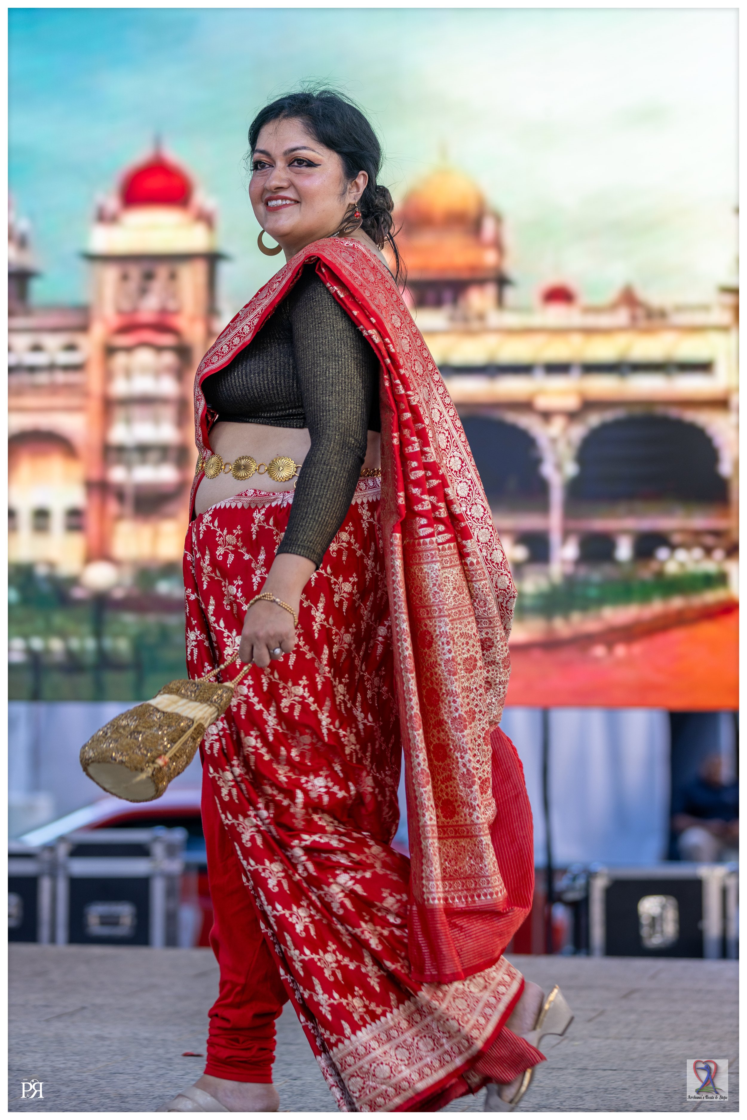 A woman smiling and dressed in traditional Indian attire, standing in front of a colorful backdrop that resembles a palace.