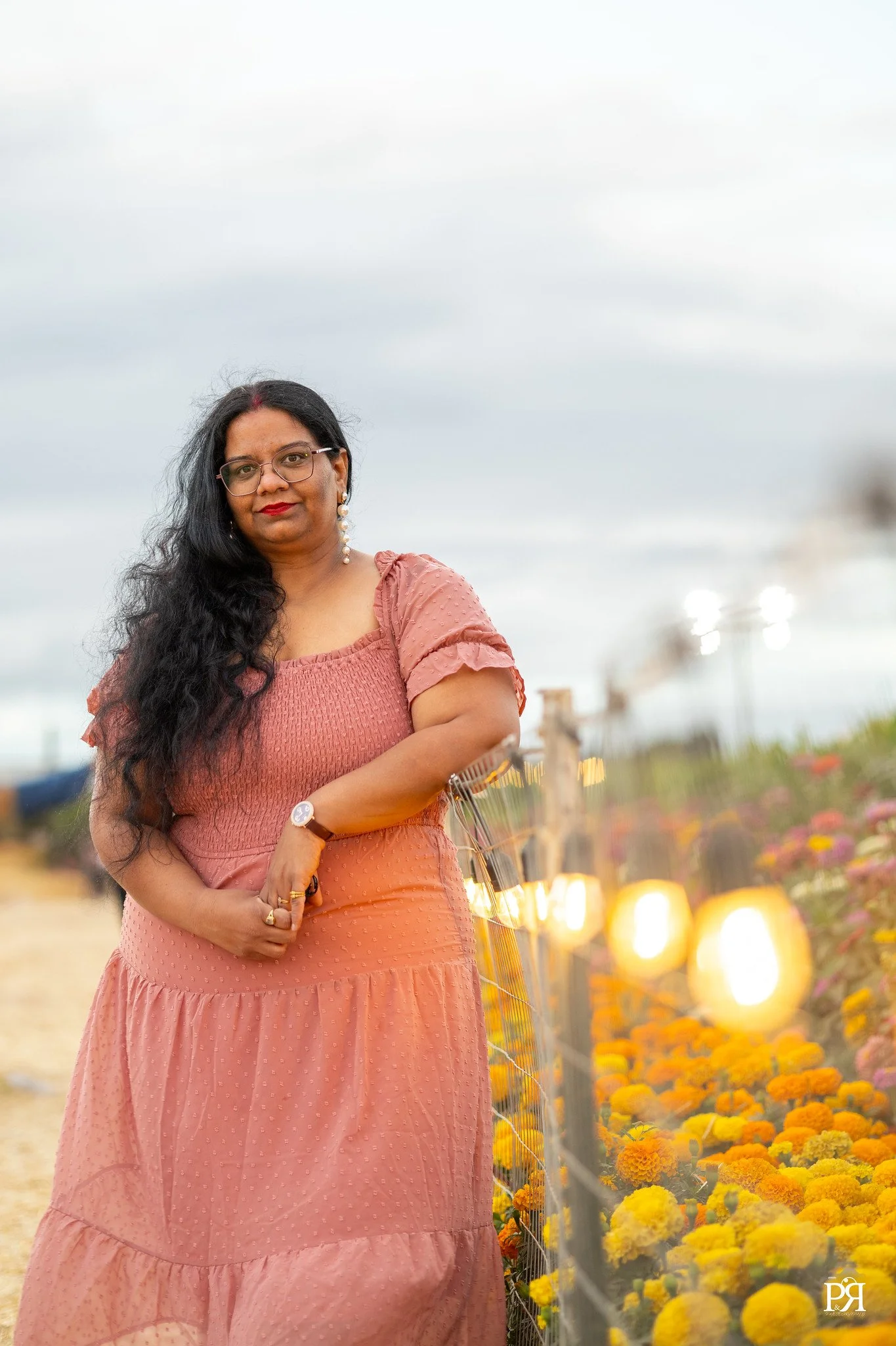 A woman in a pink dress stands near a flower patch with lights along a fence, with cloudy sky in the background.