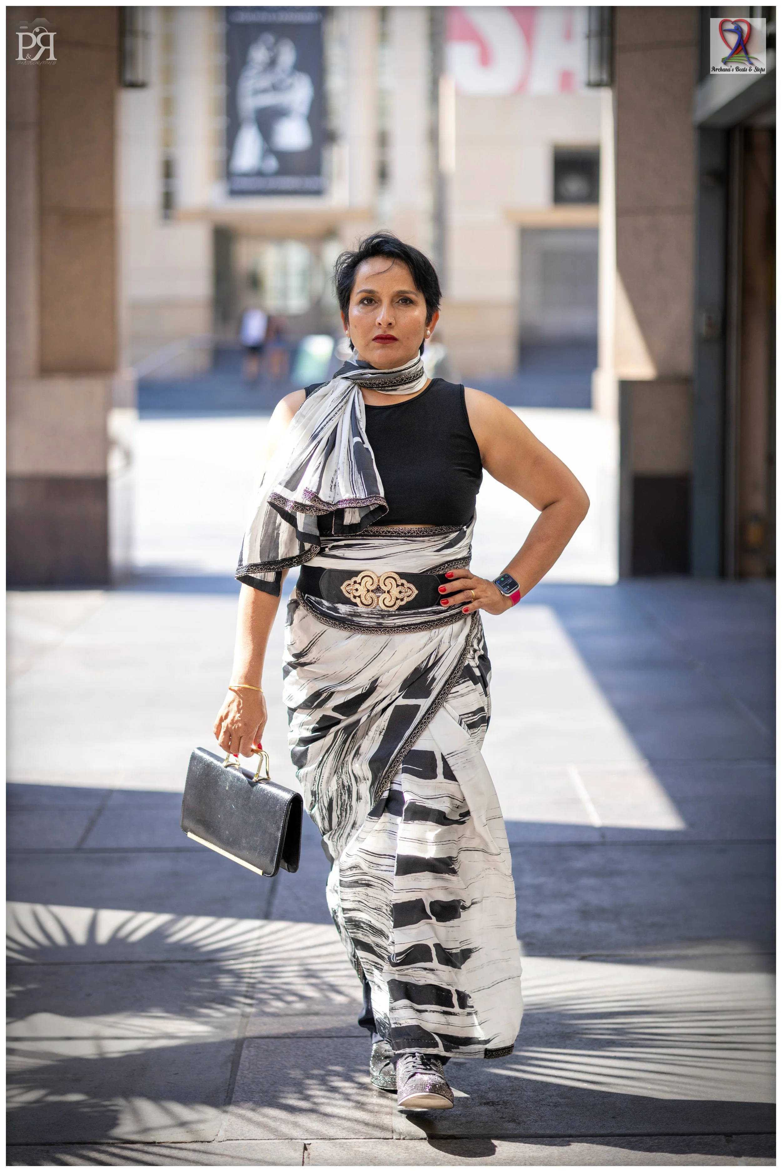 A woman walking outdoors, wearing a black top and a black, white, and gray patterned skirt and scarf, carrying a black purse, with a smartwatch on her wrist, and wearing sneakers.