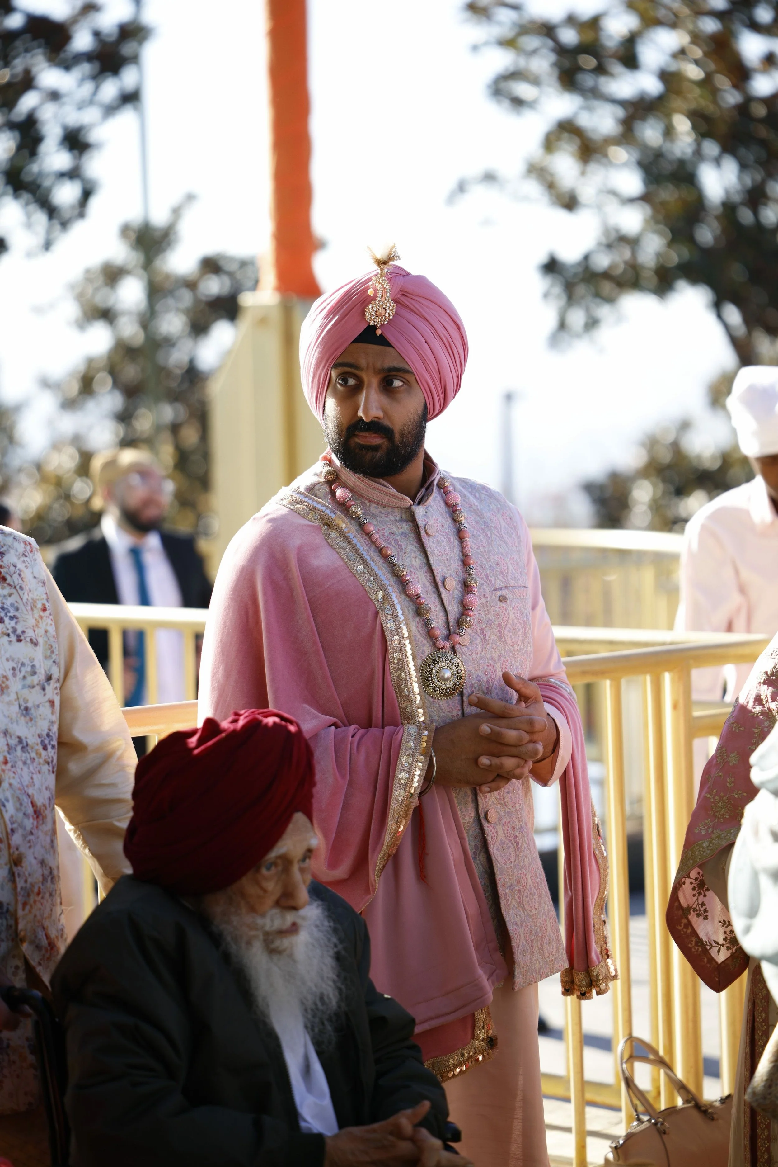 A man dressed in traditional Indian attire, wearing a pink turban and jewelry, standing with his hands clasped. An elderly man in a red turban and black outfit is seated nearby, and other people are visible in the background, outdoors with trees and 