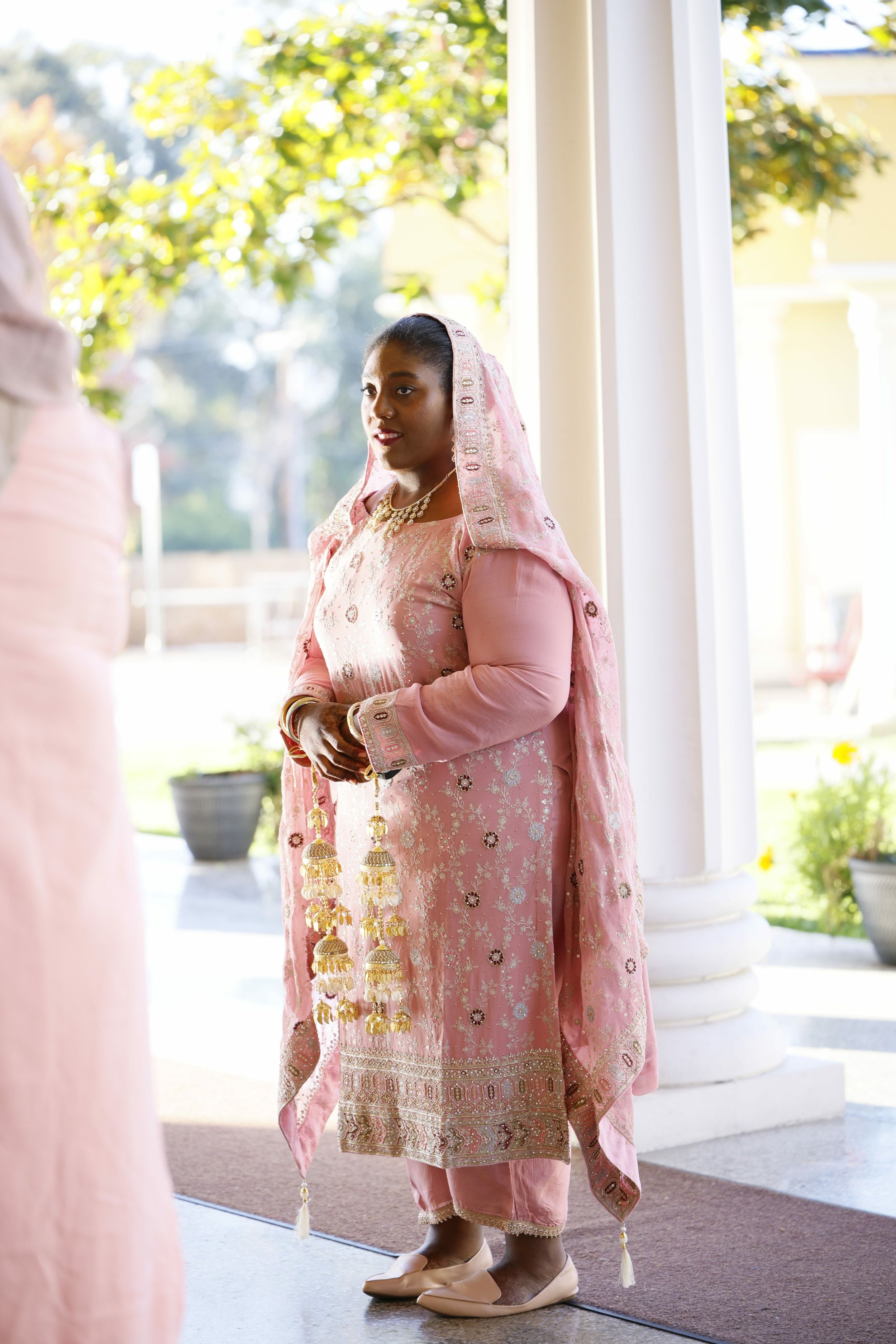 A woman dressed in traditional pink Indian attire standing near a white column, holding decorative hanging ornaments, outside with greenery in the background.
