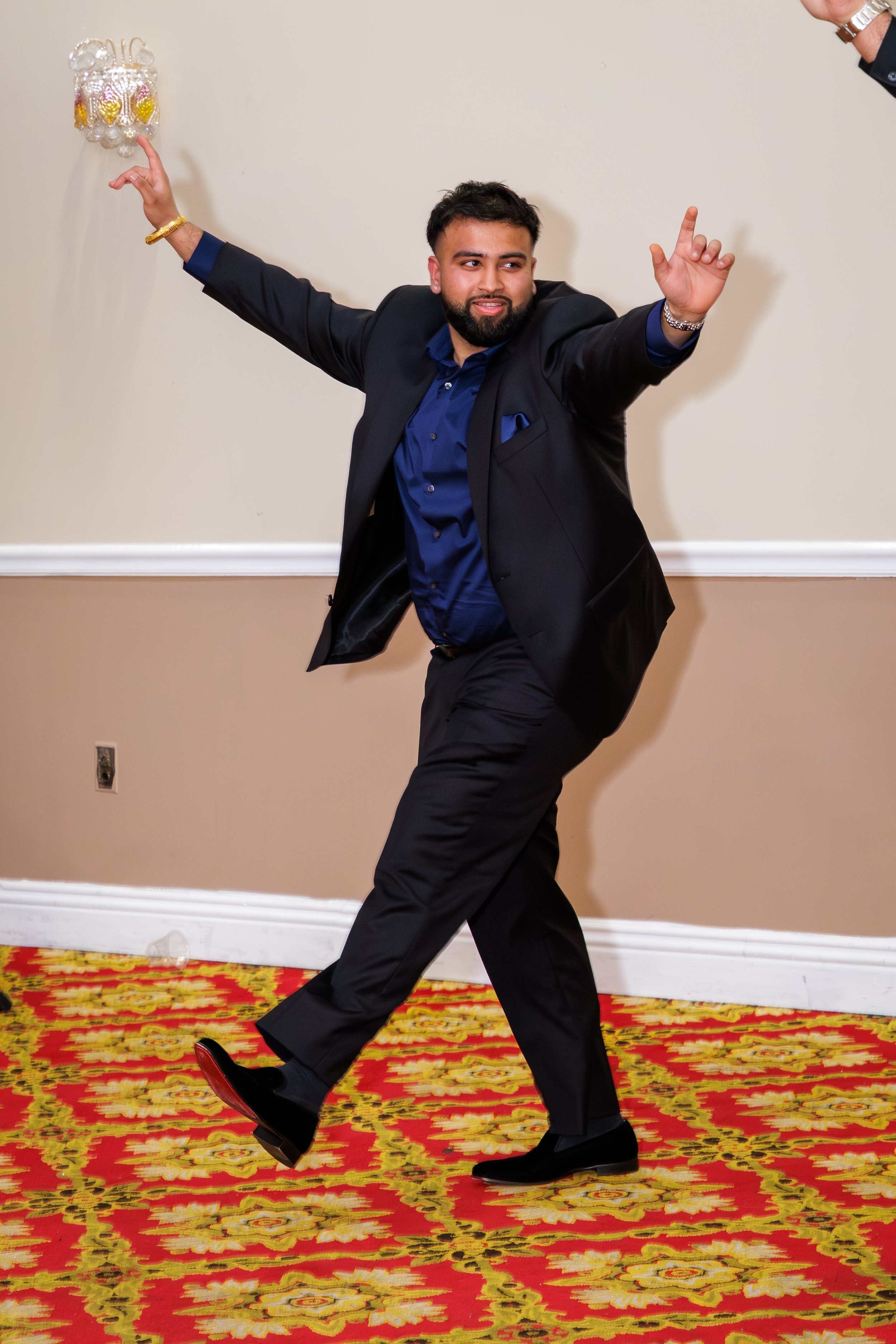 A man in a dark suit and blue shirt dancing on a patterned red and yellow carpet at an indoor event.