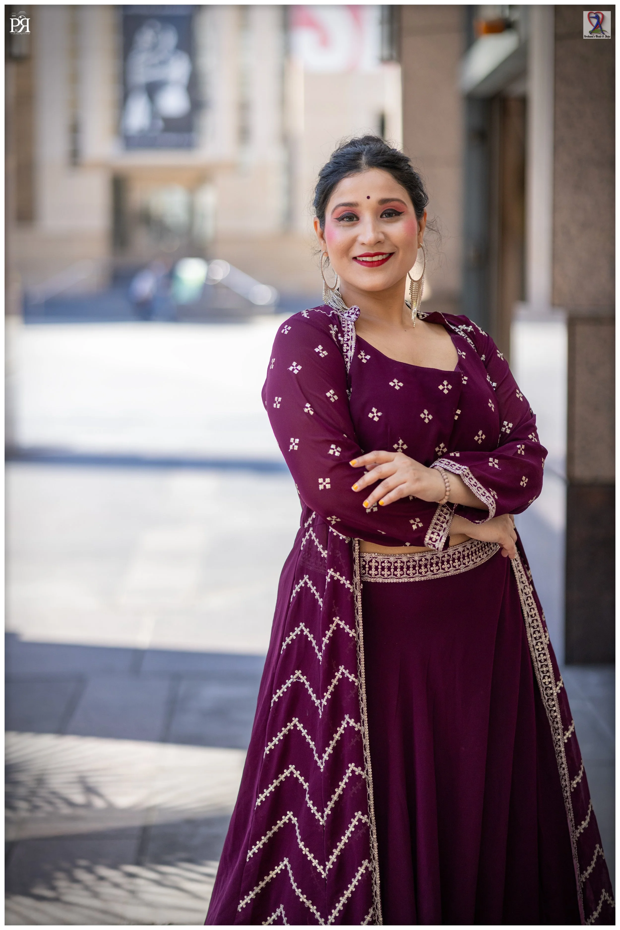 A woman in traditional Indian attire, with a purple dress with white embroidery, standing outdoors with buildings in the background, smiling and crossing her arms.