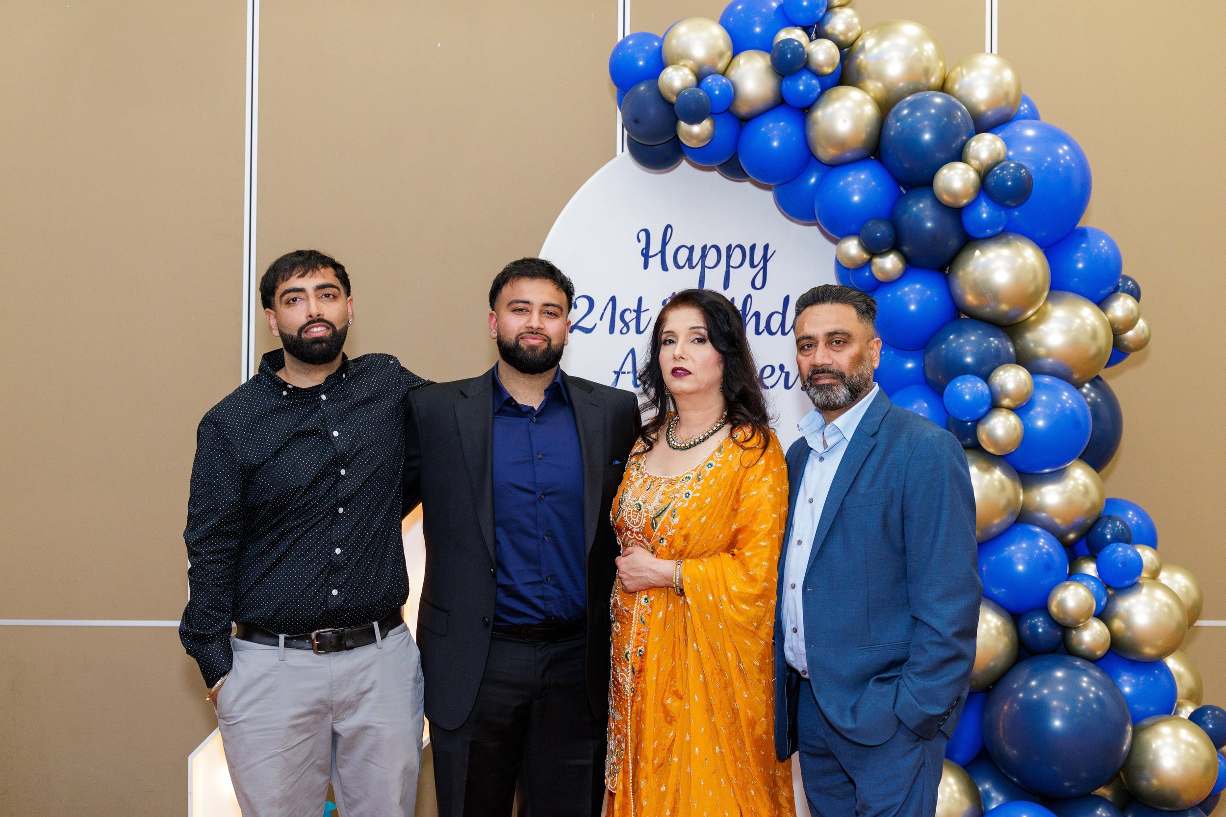 Four people standing in front of a balloon arch with gold and blue balloons, celebrating a 21st birthday with a sign that says 'Happy 21st Birthday A...'.