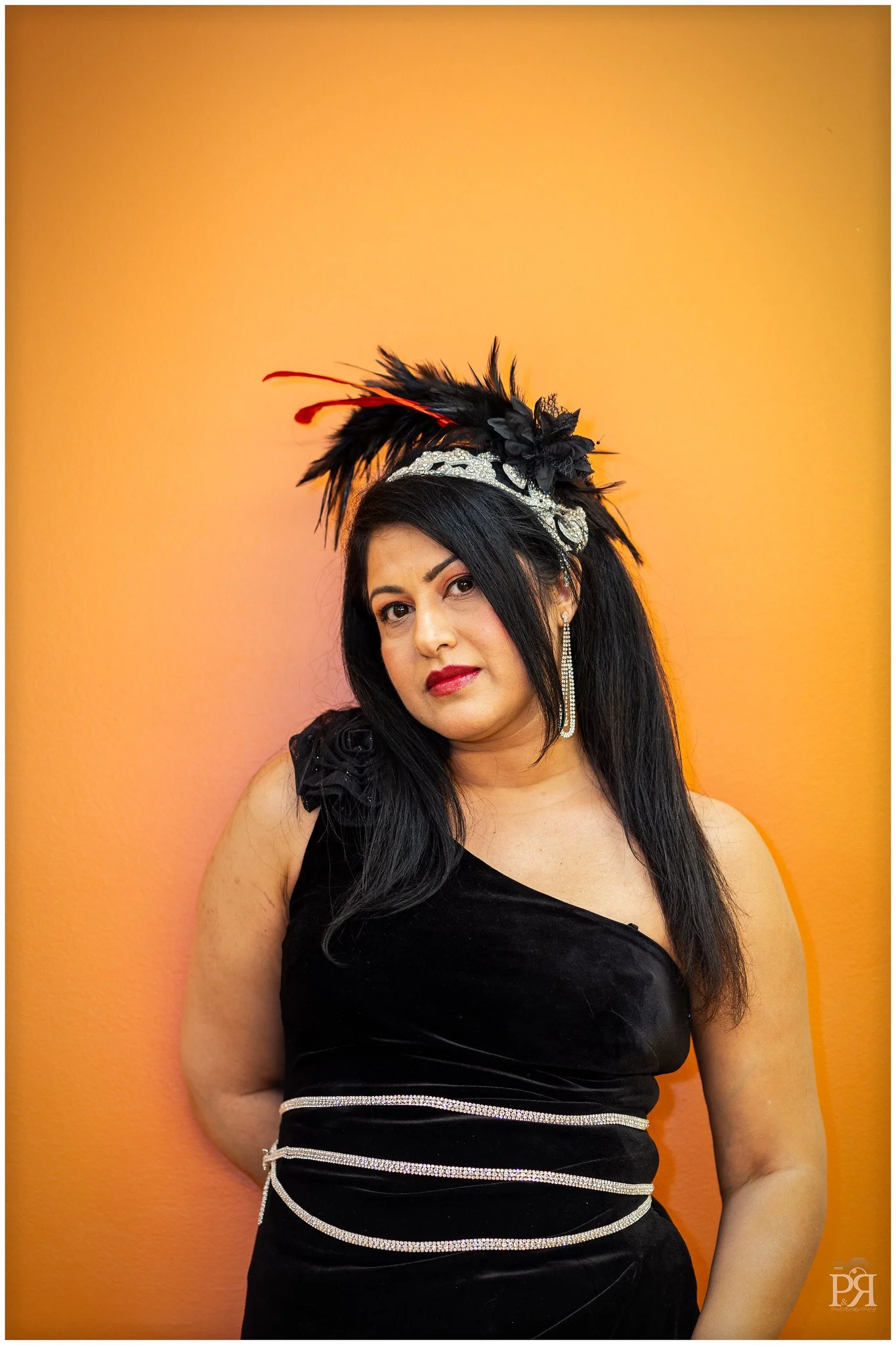 A woman with dark hair in a black dress with rhinestone accents, wearing a feathered headpiece and long earrings, standing against an orange wall.