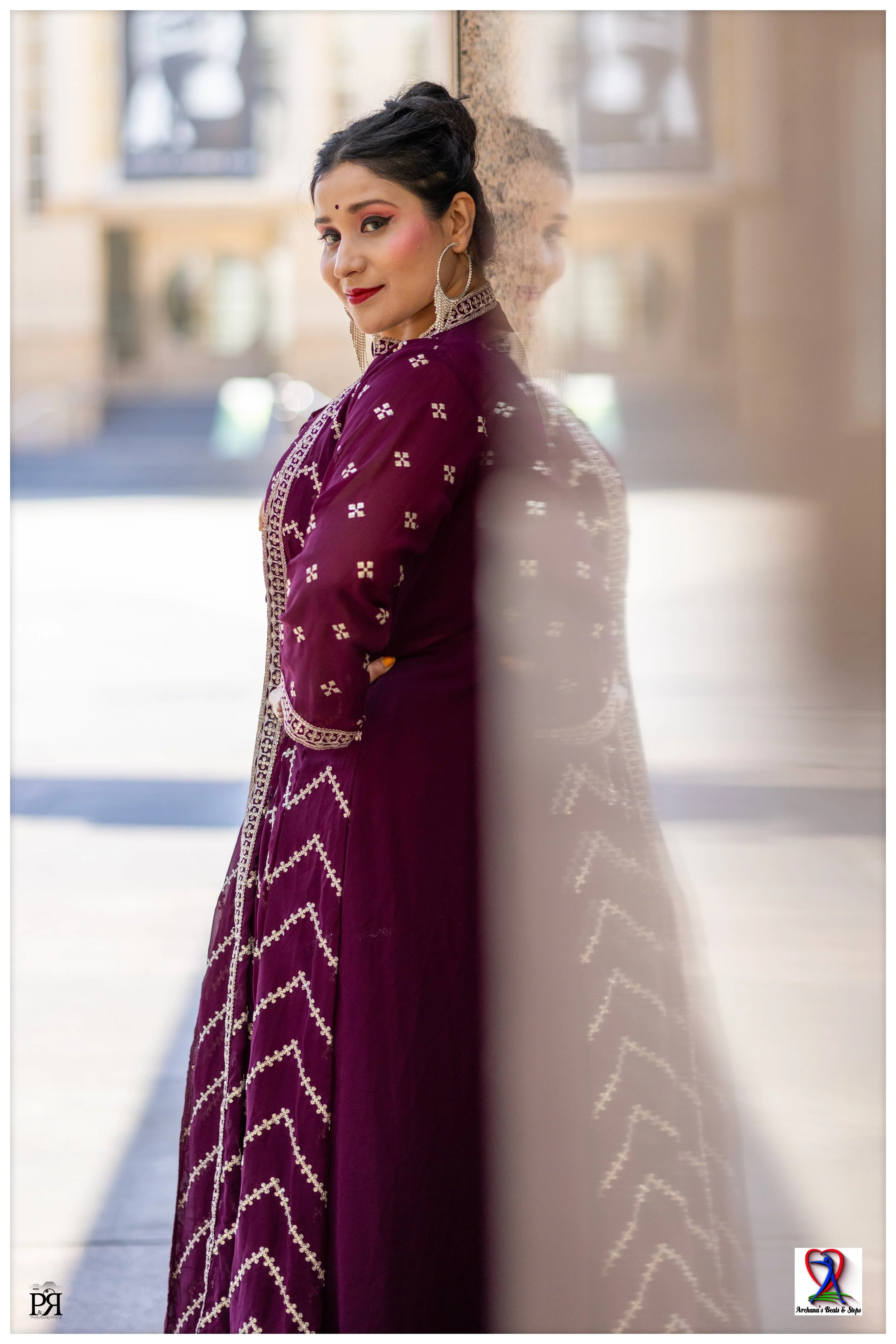 A woman in traditional Indian attire, wearing a deep purple embroidered dress, stands by a glass wall with her reflection visible on it, outdoors with blurred buildings in the background.