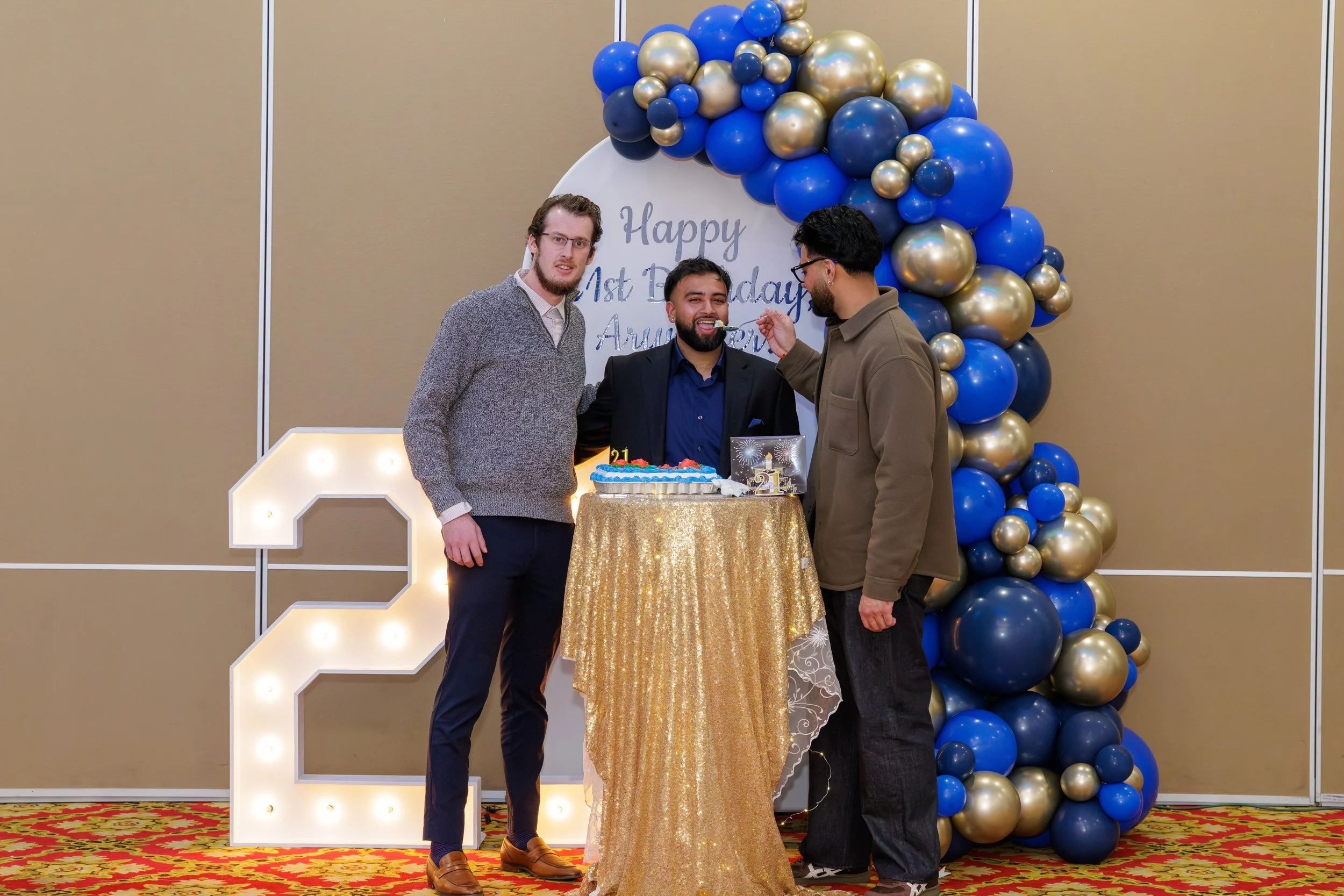 Three men celebrating a birthday at a decorated party. One man is blowing out a candle on a birthday cake, while the other two stand beside him. They are in front of a backdrop decorated with blue and gold balloons and a sign that says 'Happy 1st Bir