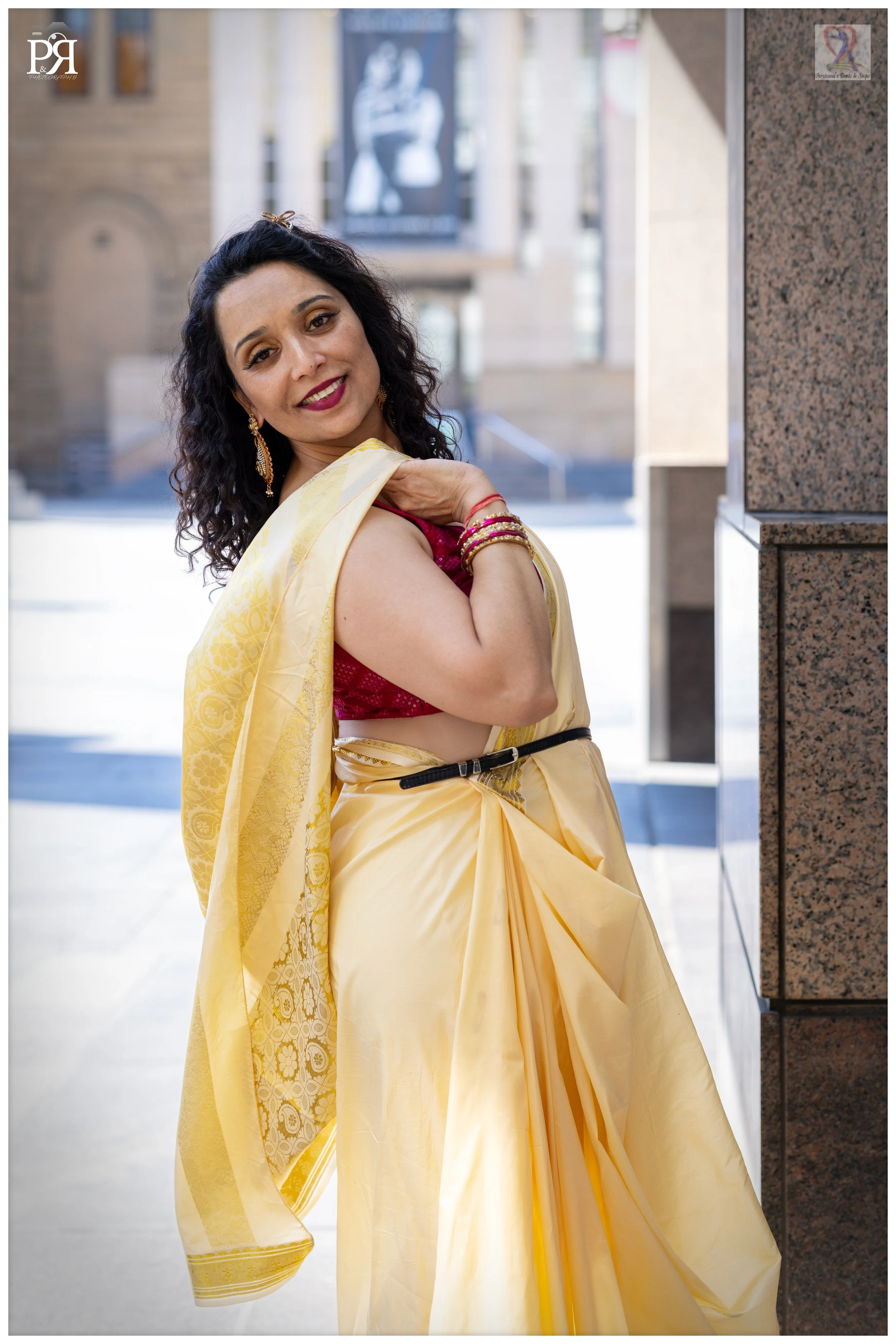 Woman dressed in a yellow sari with a red blouse, standing outdoors, smiling, with architectural buildings in the background.