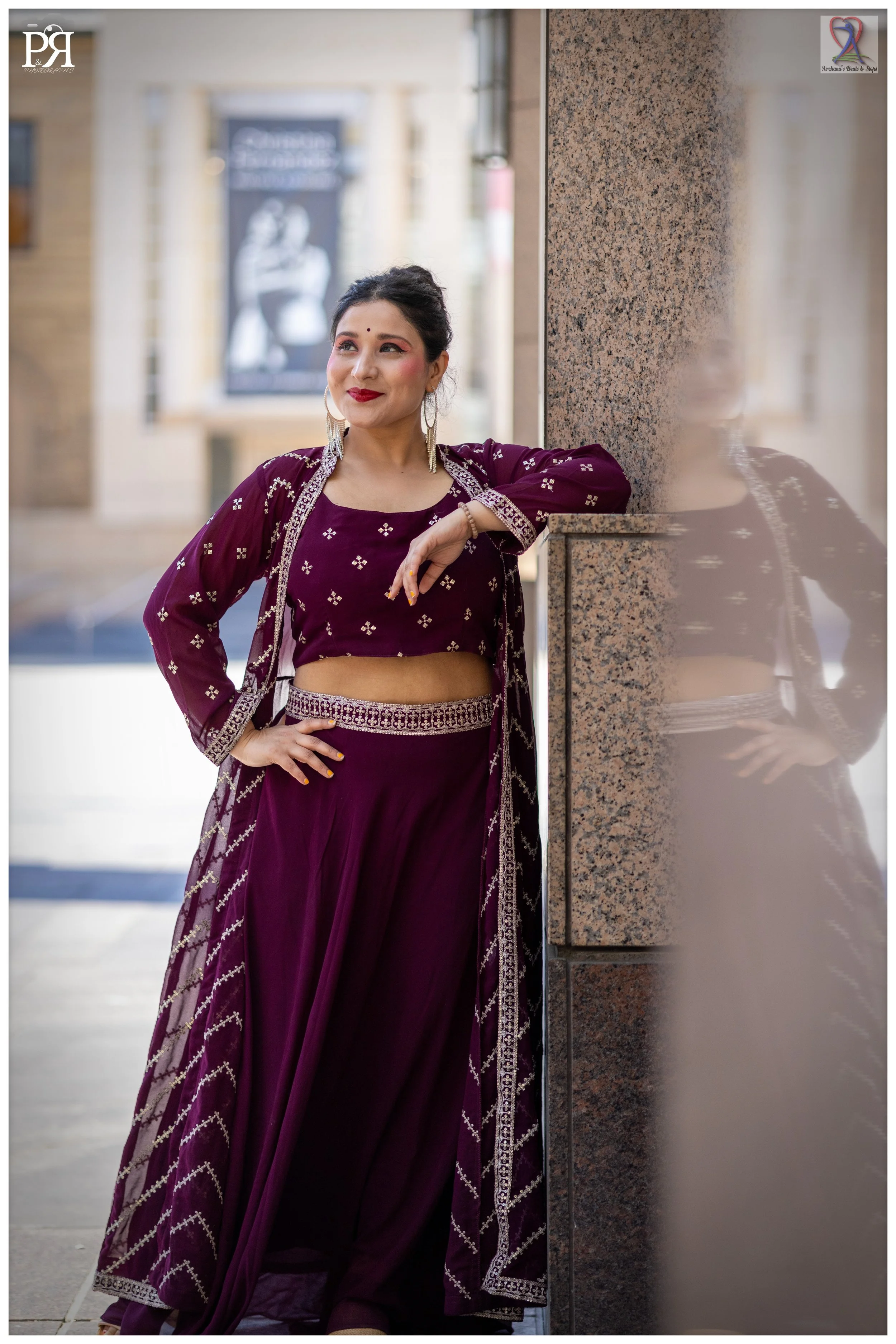 A woman in a purple traditional Indian outfit, standing outdoors with her arm resting on a stone wall, smiling and looking to her left, with her reflection visible on the wall.