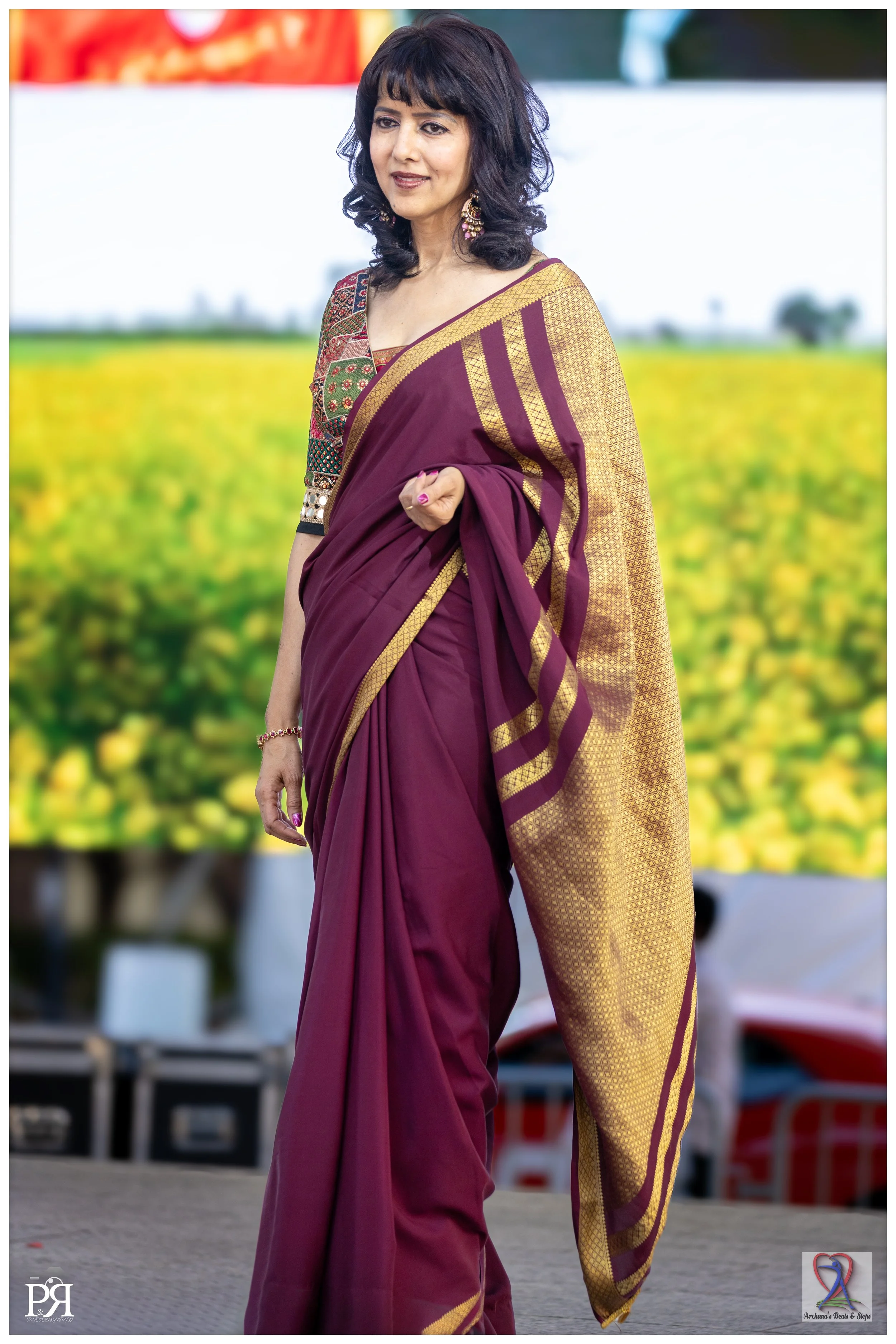 A woman in a maroon and gold sari standing outdoors in front of a yellow flower field, with trees and a blurred background.