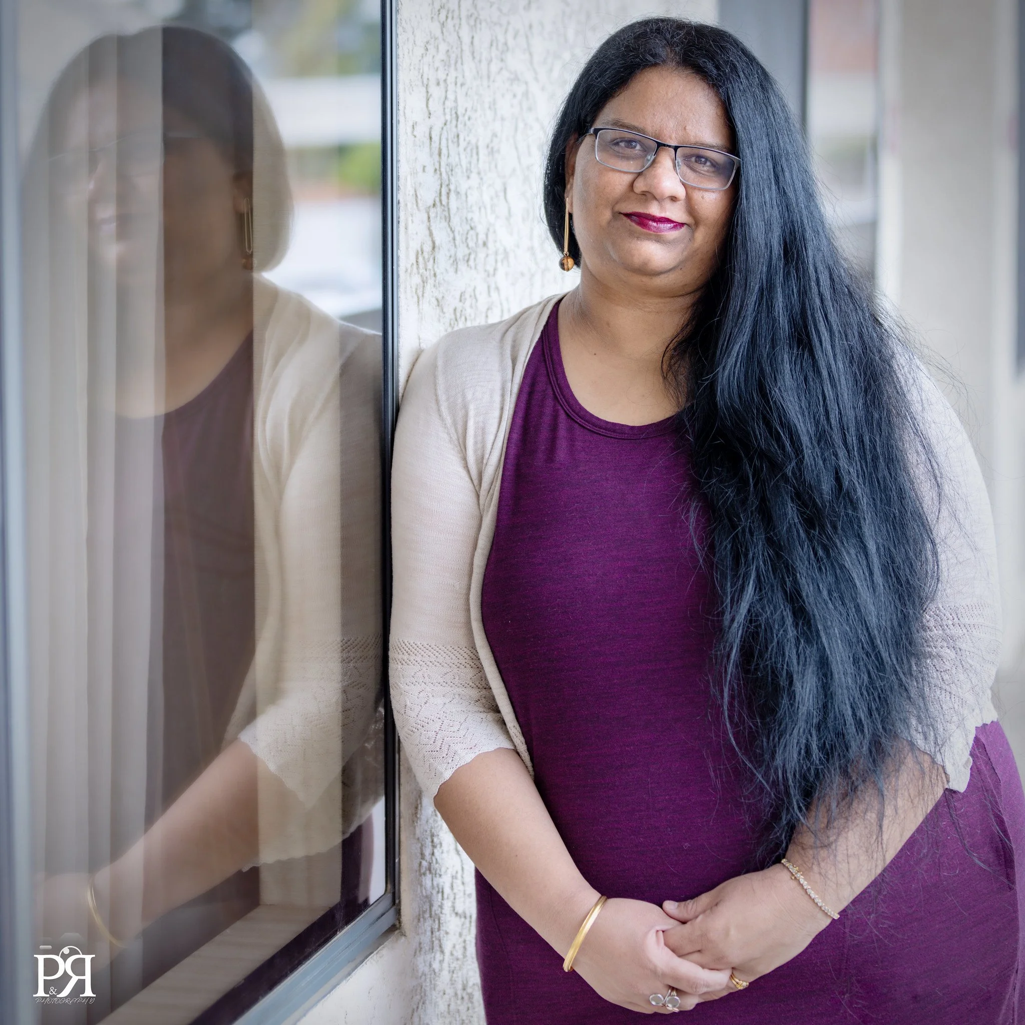 A woman with long black hair, glasses, and a purple dress stands beside a window, smiling.