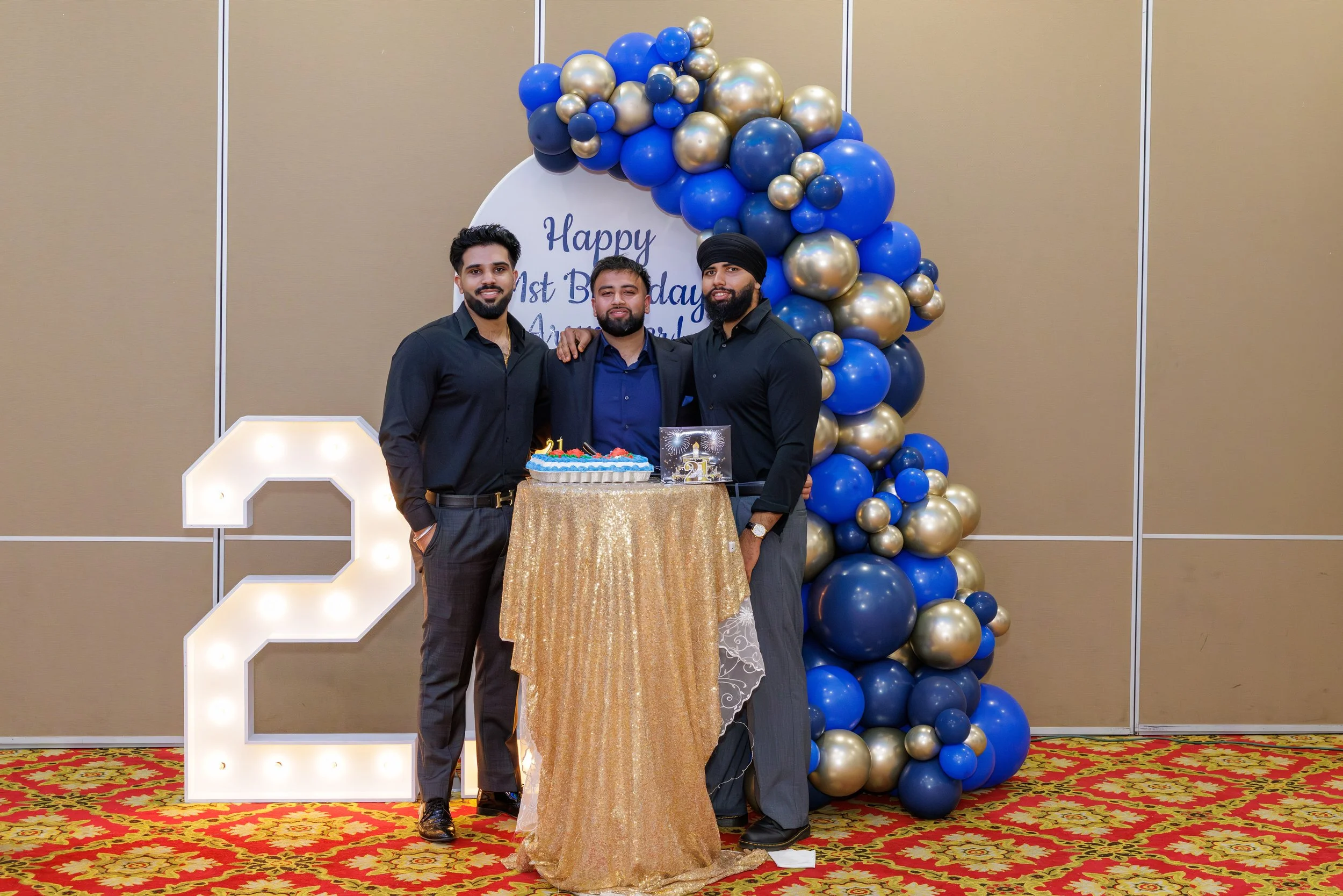 Three men celebrating a birthday with a cake in front of a large balloon arch and a lit-up number 2. The background features a sign that says 'Happy 1st Birthday.' The floor has a red and gold patterned carpet.