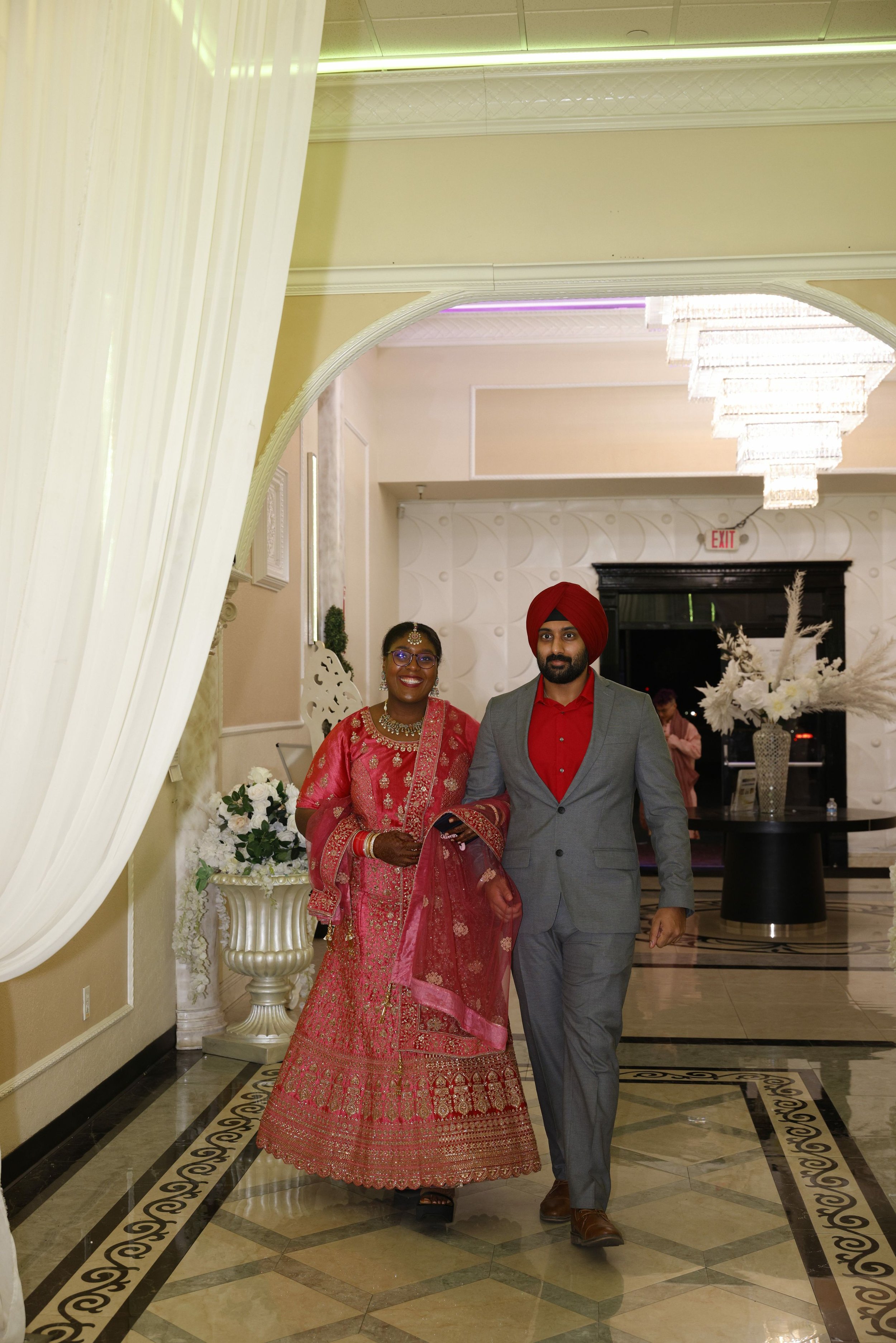 A couple dressed in traditional South Asian wedding attire, walking together in an elegantly decorated indoor venue with marble floors, white floral arrangements, and a large chandelier.