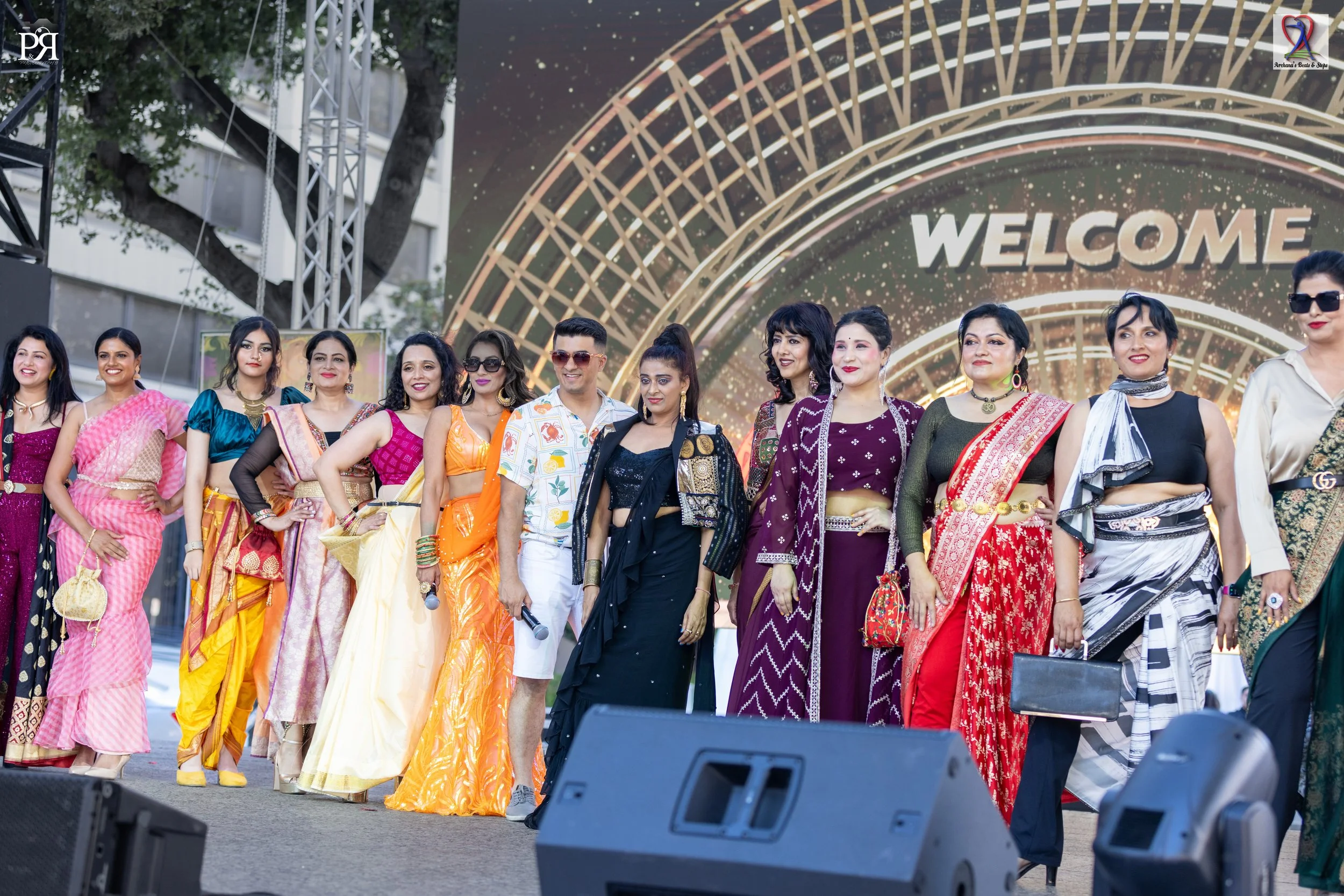A group of people standing on stage in front of a large digital backdrop displaying the word 'WELCOME' and colorful geometric designs, dressed in traditional and modern clothing.