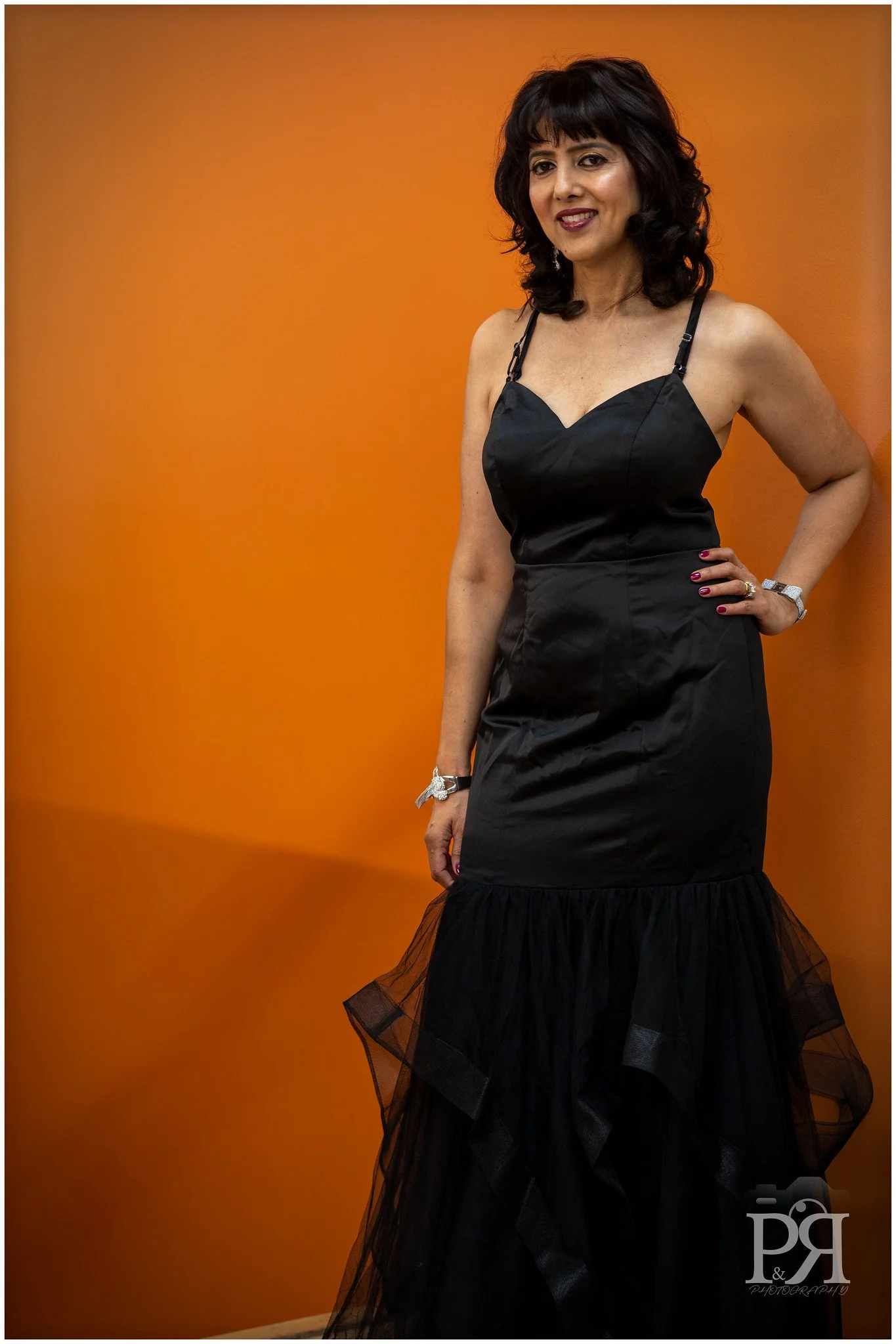 A woman with dark, curly hair wearing a black dress with spaghetti straps, standing against an orange background, smiling with her left hand on her hip.