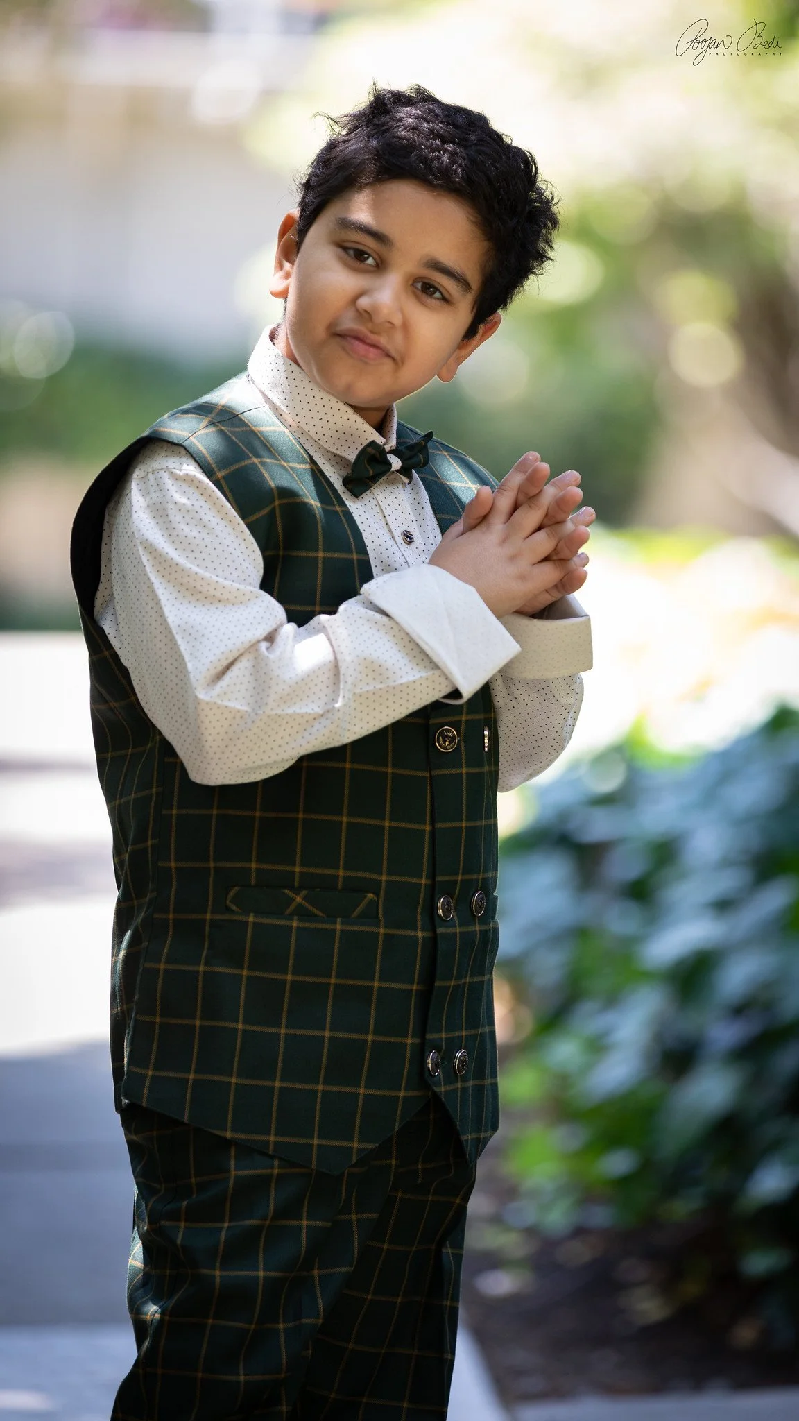 A young boy with dark curly hair in formal attire, standing outdoors with hands clasped together, looking at the camera with a slight smile.