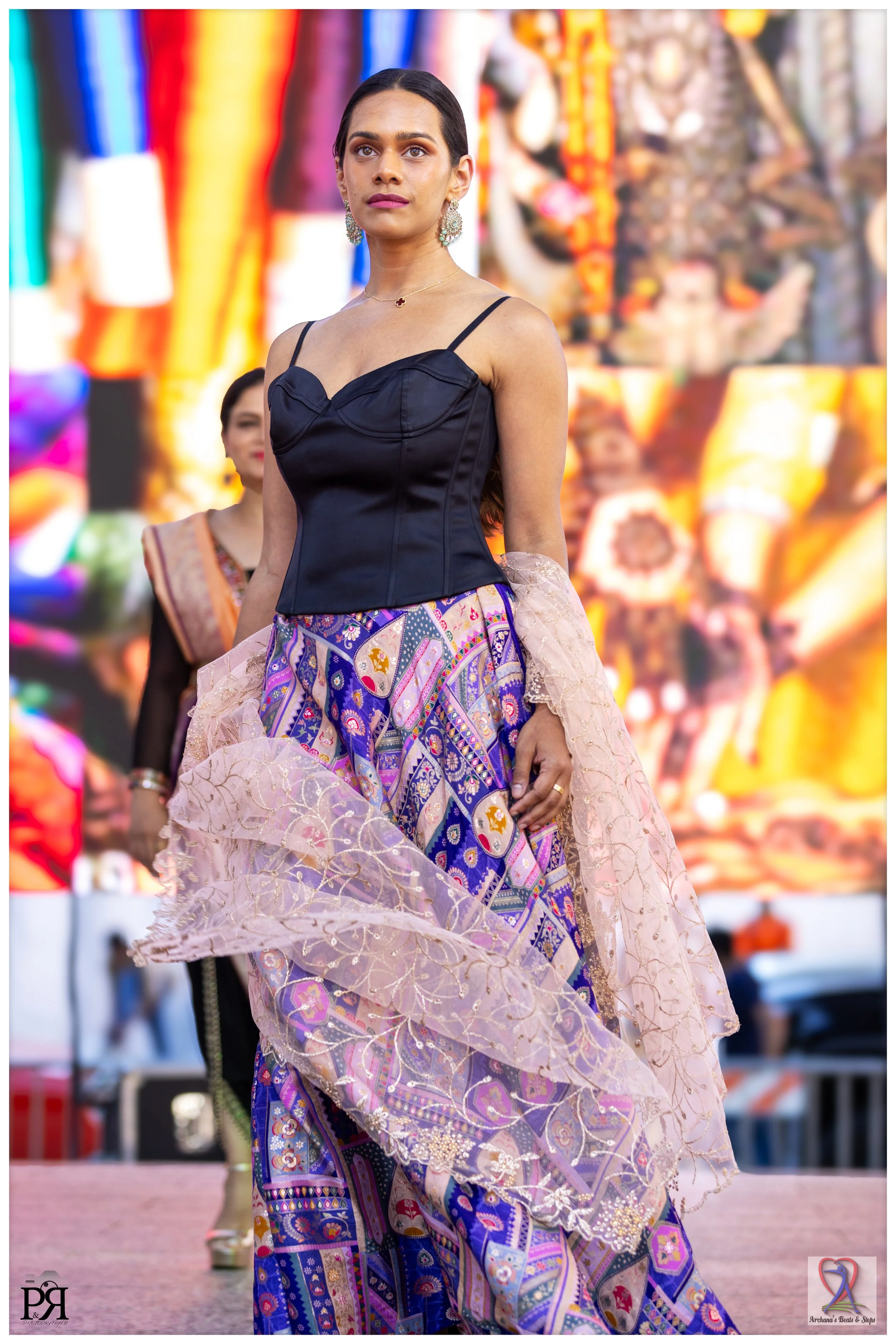 A woman walking on stage in a black top and colorful, patterned skirt, with a semi-transparent pink shawl, at an outdoor event with a vibrant, colorful background.