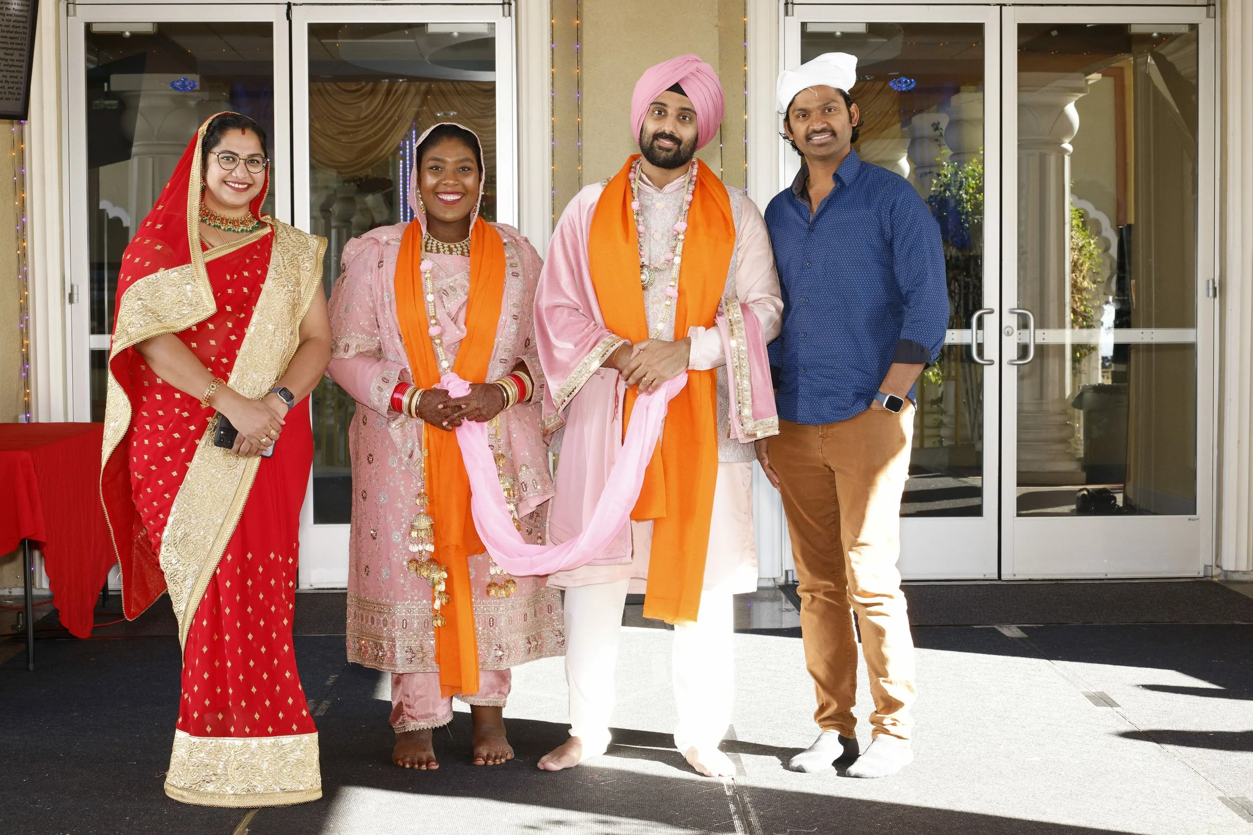 Group of five people dressed in traditional Indian attire standing in front of glass doors at a celebration or wedding event.