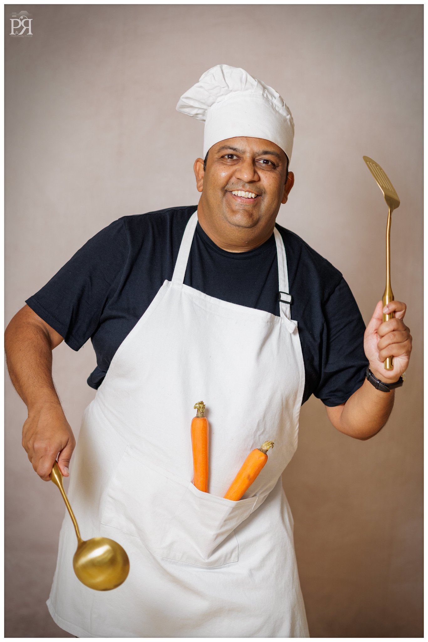 A smiling man dressed as a chef wearing a white chef hat and apron, with three carrots tucked into his apron pocket. He is holding a gold cooking spoon in his right hand and a gold spatula in his left hand, standing against a neutral background.