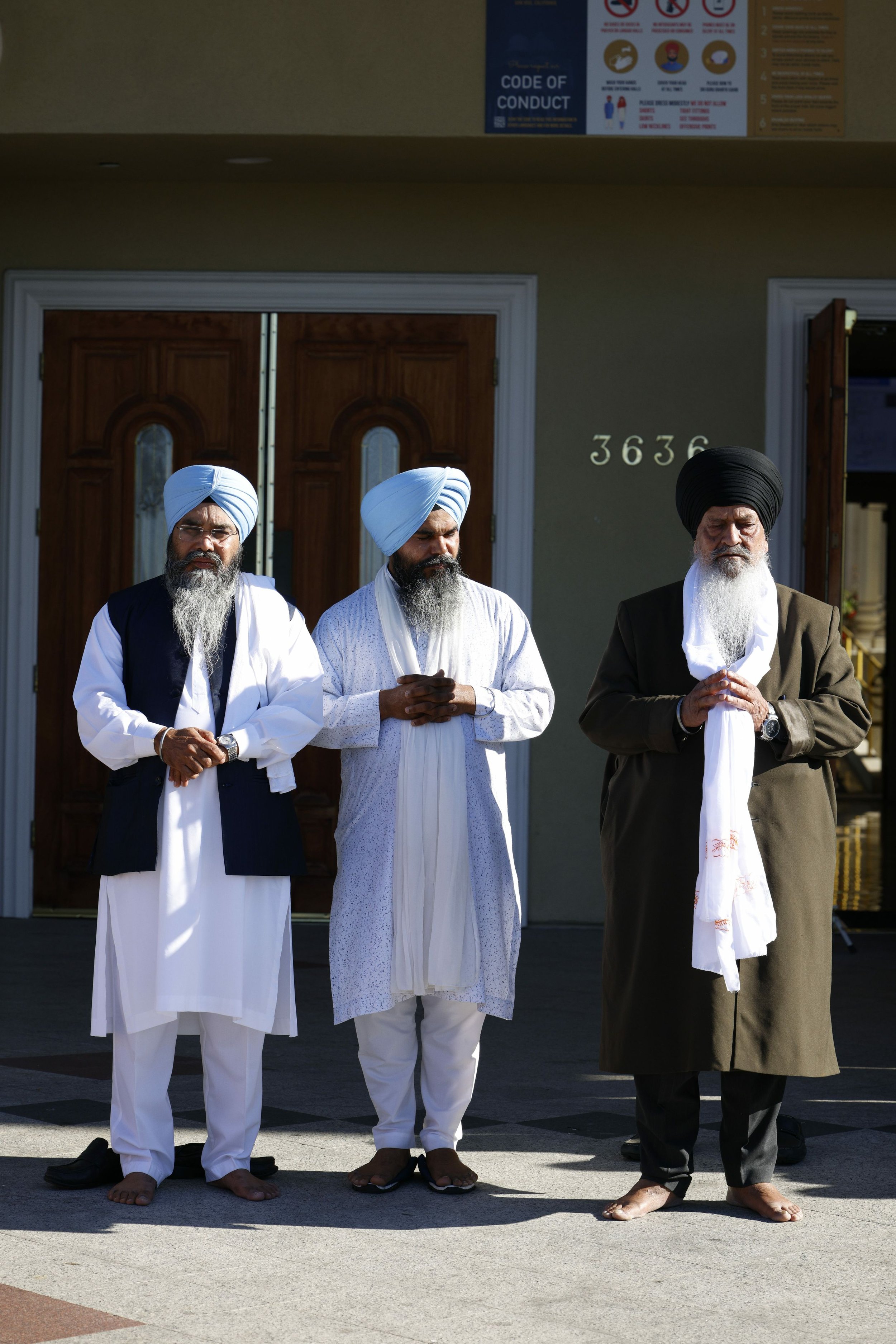 Three Sikh men wearing turbans and traditional attire standing with hands clasped in prayer outside a building.