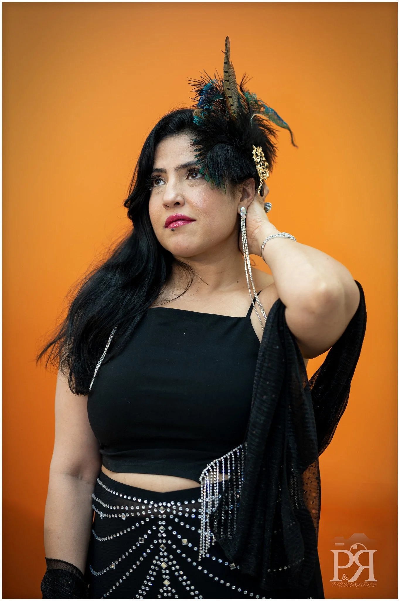 A woman with long black hair wearing a black outfit with rhinestones and jewelry, holding an ornate feathered headdress, against an orange background.