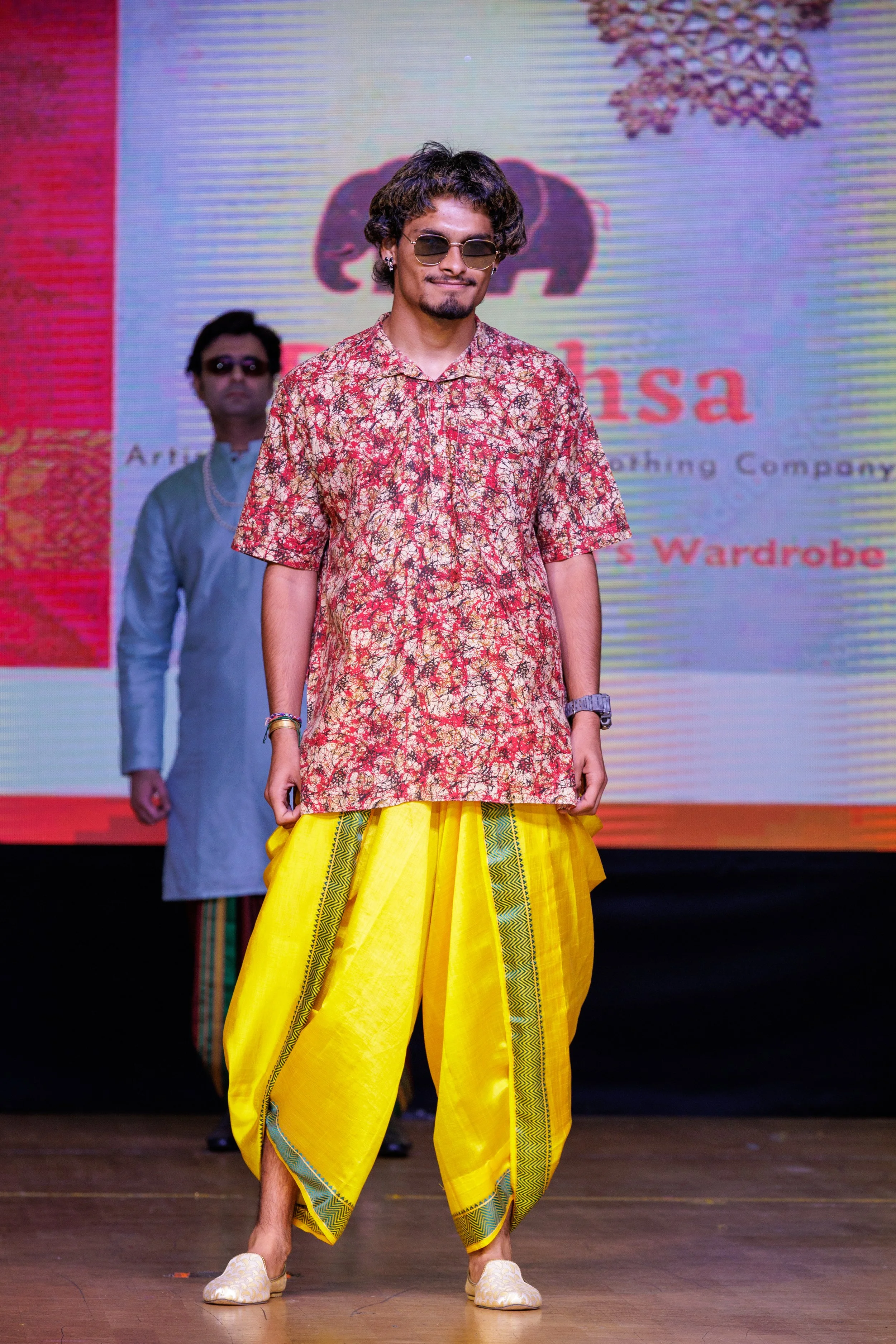 A male model walking on stage wearing a traditional Indian outfit with a red patterned kurta and bright yellow dhoti pants, sunglasses, and traditional footwear, with a large screen in the background displaying logo and text.