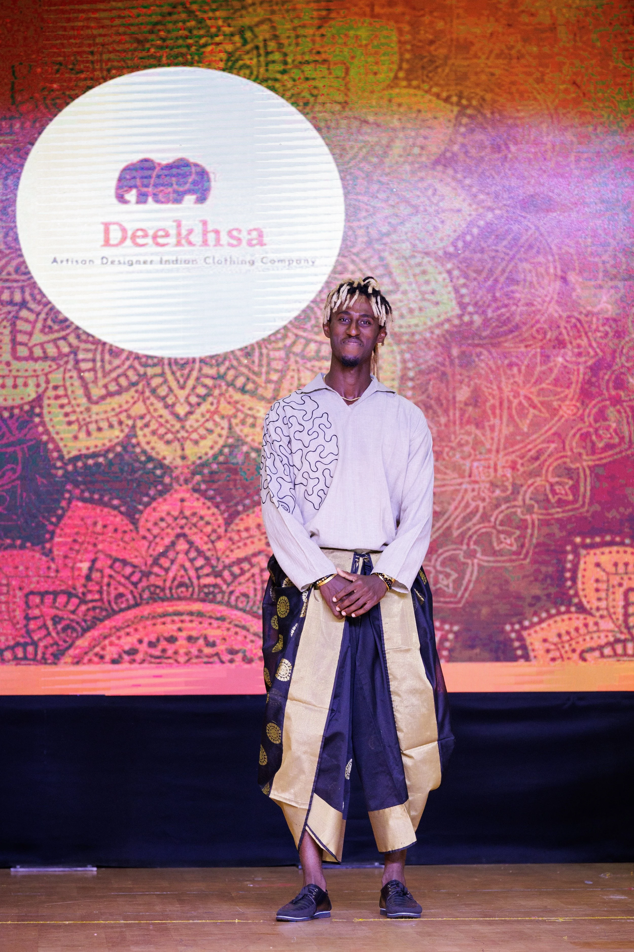 A young man stands on a stage with a colorful, mandala-like background. He is wearing a white shirt with black decorative patterns on the sleeves and beige and black traditional African-style pants. Behind him, a large screen displays a logo with a p