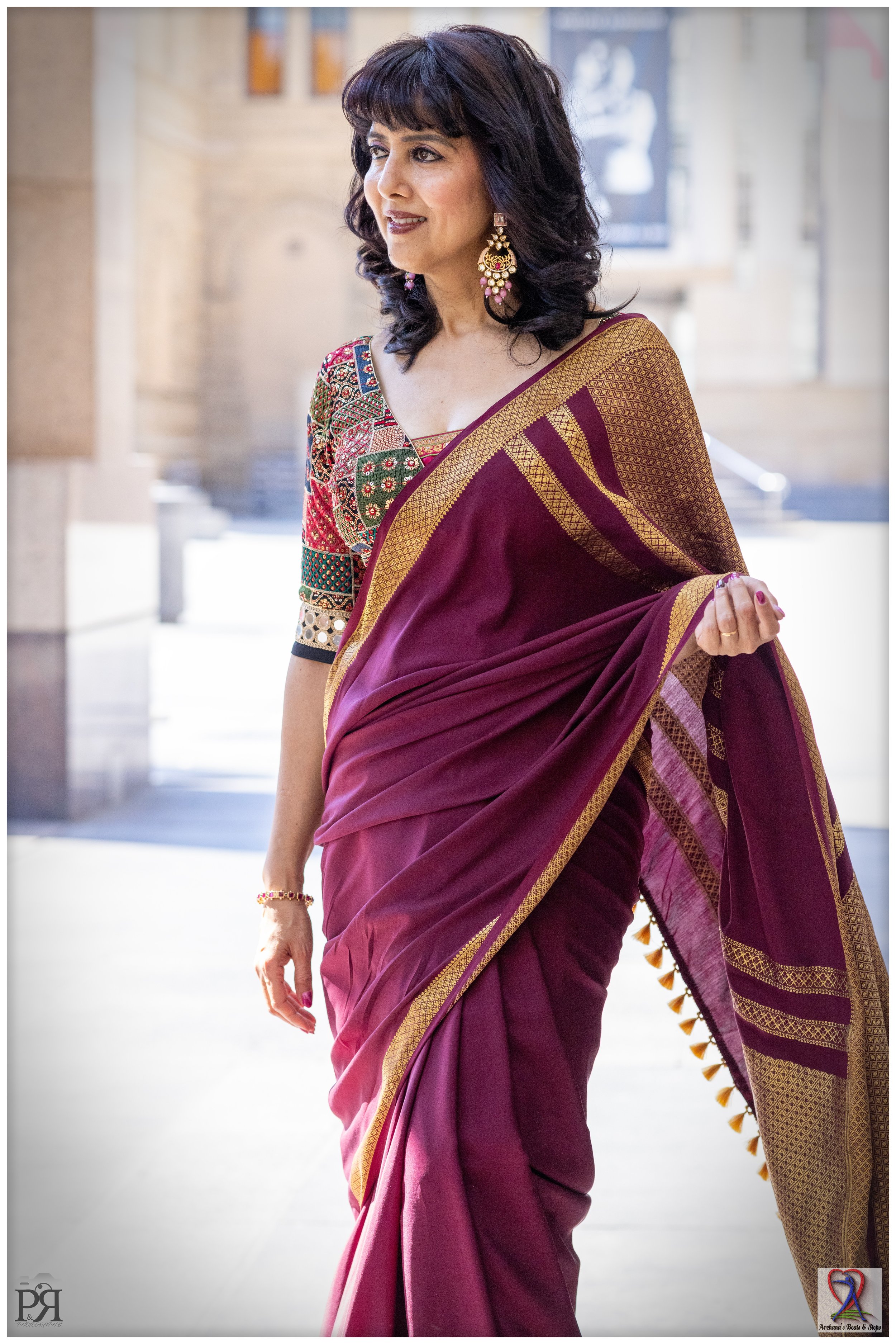 A woman in traditional Indian attire, wearing a maroon saree with gold accents and a multicolored blouse with embroidery. She has dark curly hair, large earrings, and a bracelet, standing outdoors in front of a architectural backdrop.