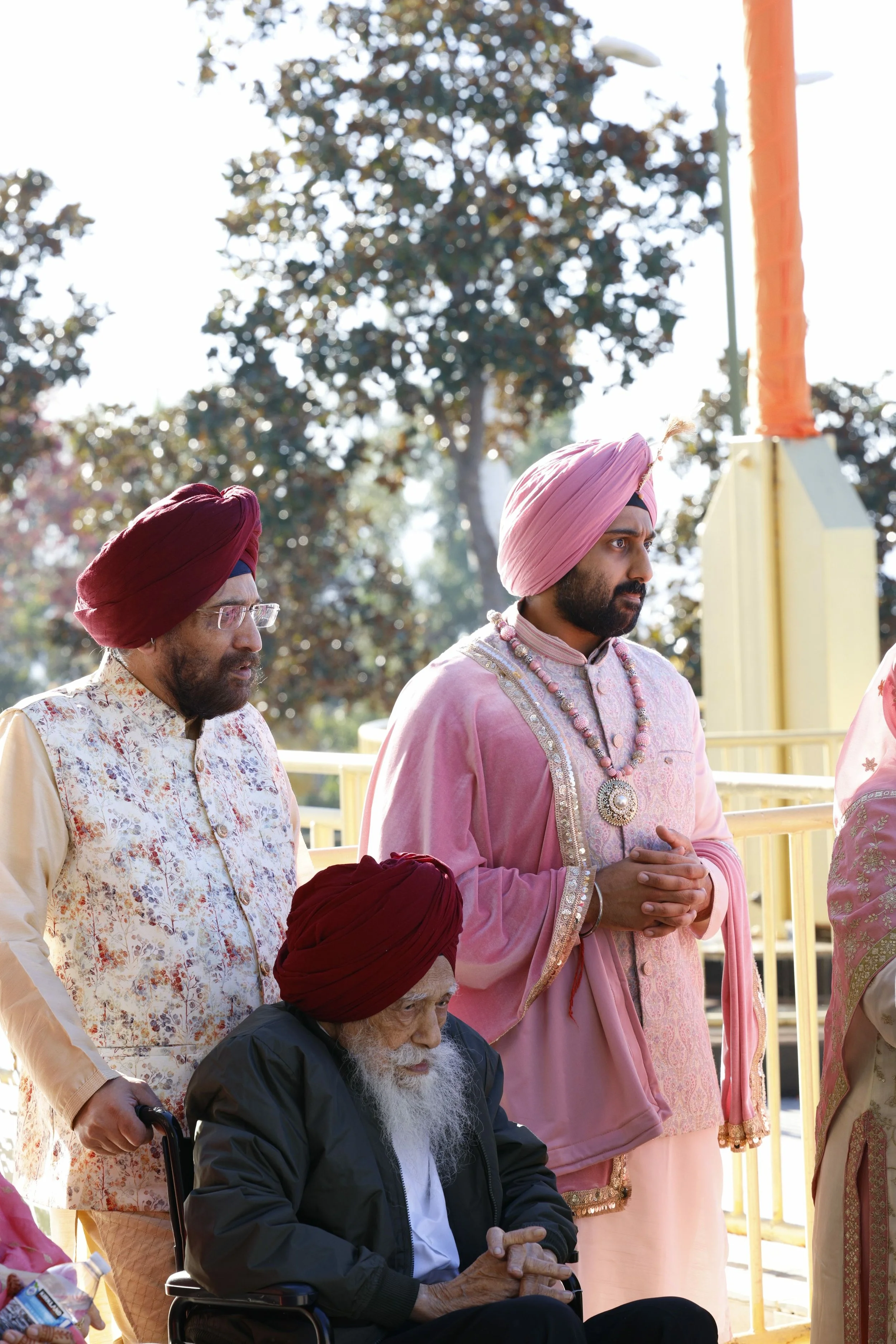 A group of Sikh men dressed in traditional attire, including turbans, participating in a religious or cultural event outdoors, with trees and a yellow railing in the background.