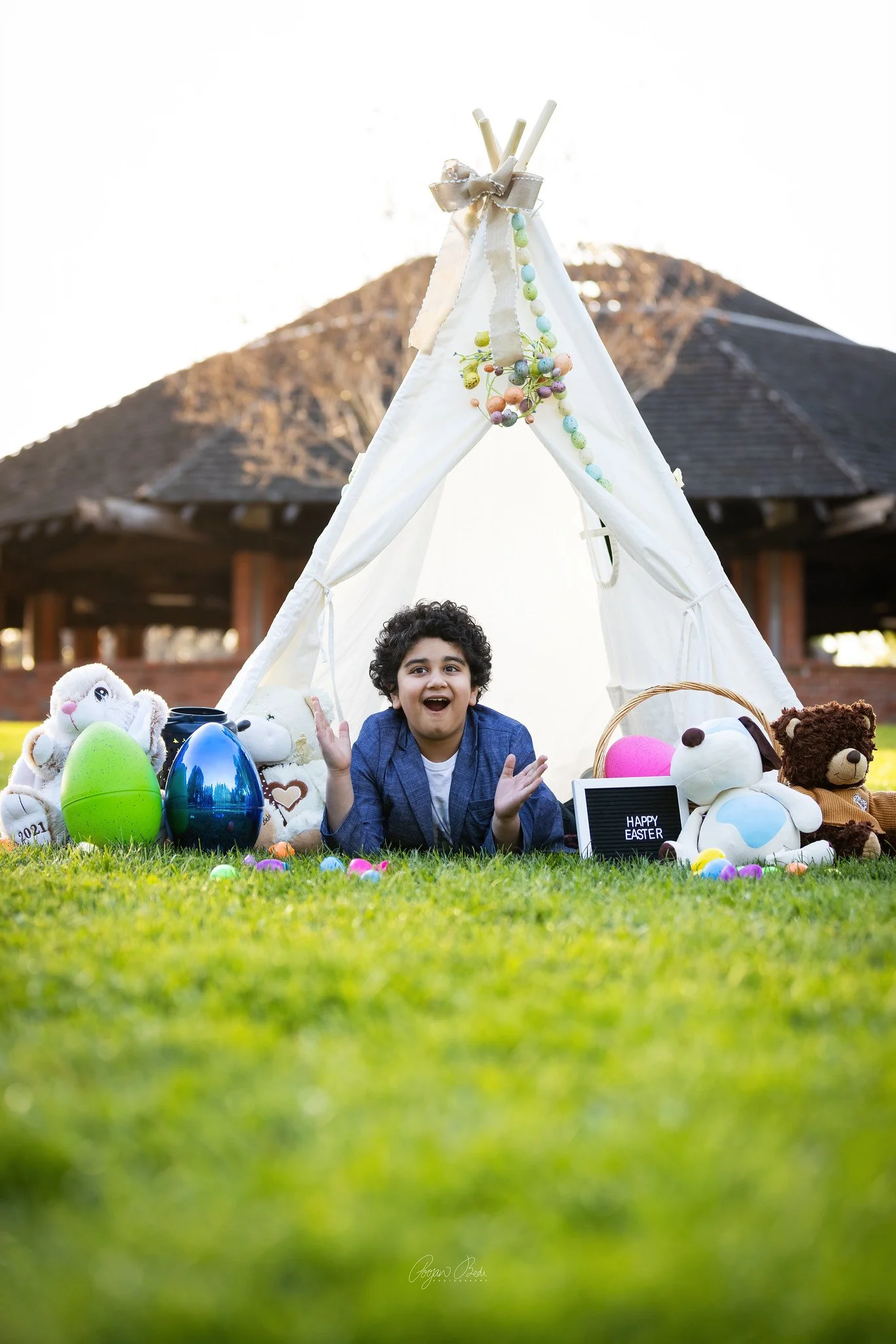 A young boy with curly hair, sitting inside a small white tent with Easter decorations, plush toys, and colorful eggs, celebrating Easter outdoors in front of a wooden pavilion.