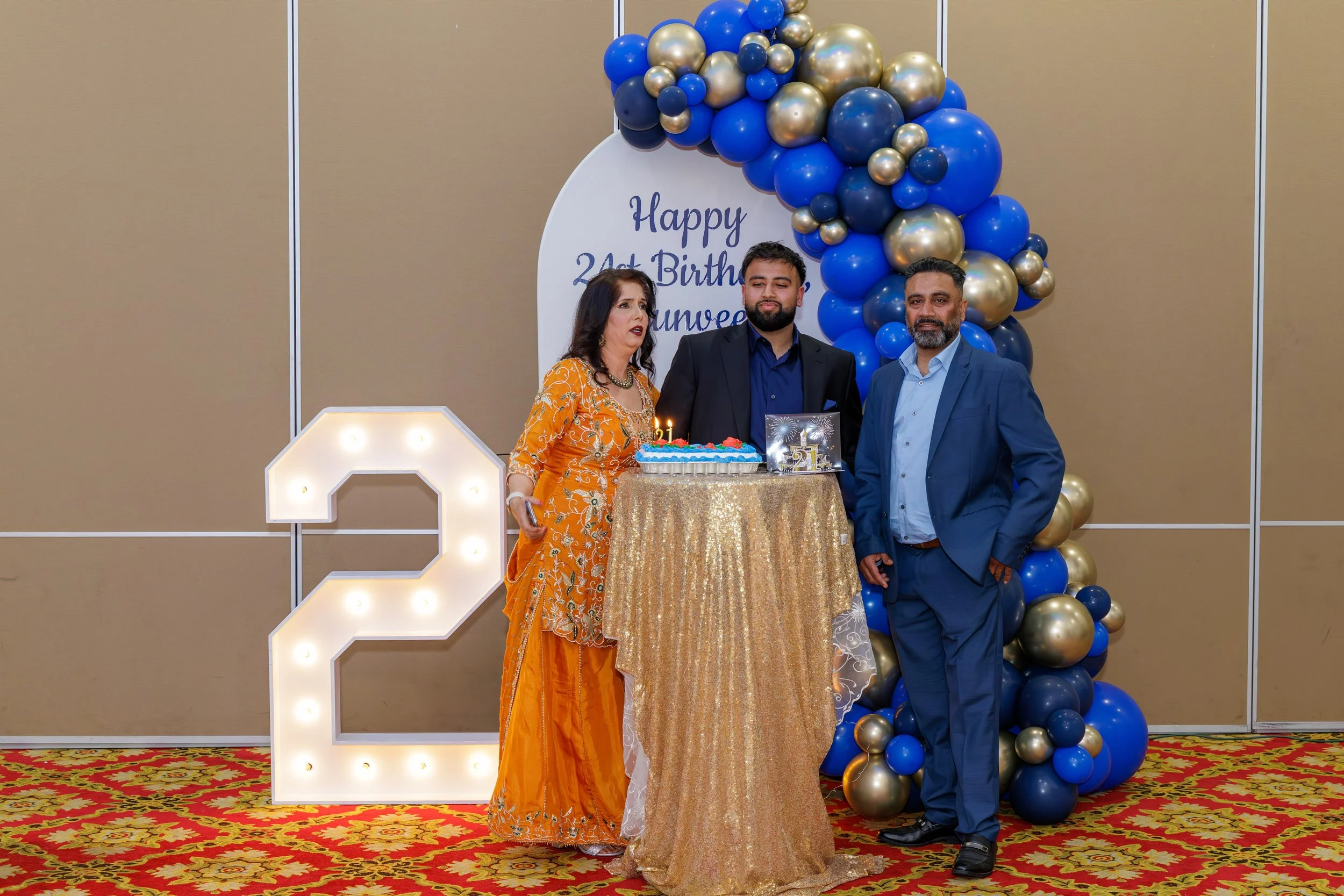 Three people standing at a birthday celebration with a decorated backdrop. A woman in an orange traditional dress stands on the left, a man in a black blazer and blue shirt in the middle, and another man in a blue suit on the right. There is a golden