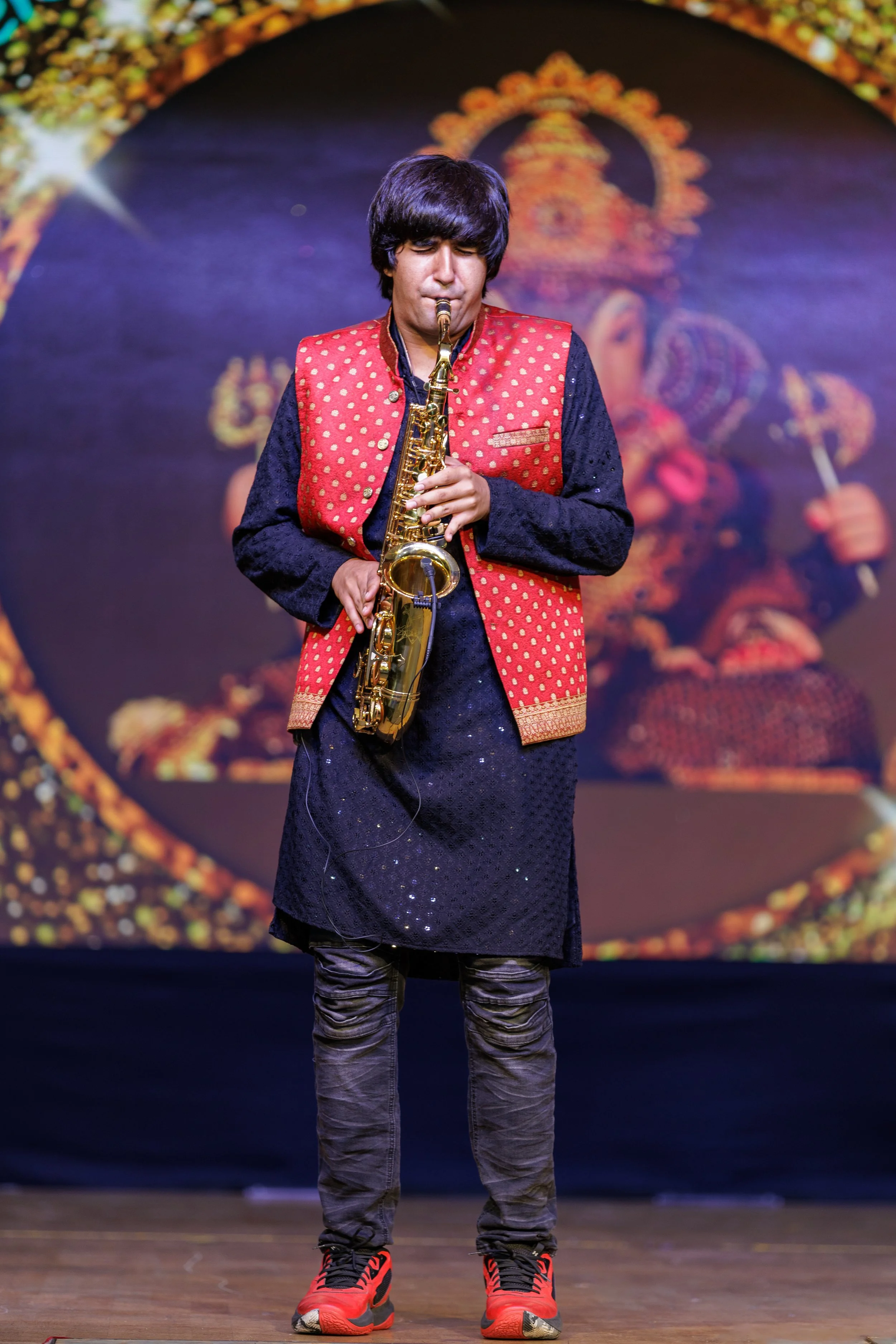 A man in traditional Indian attire playing a saxophone on stage with a decorative background.