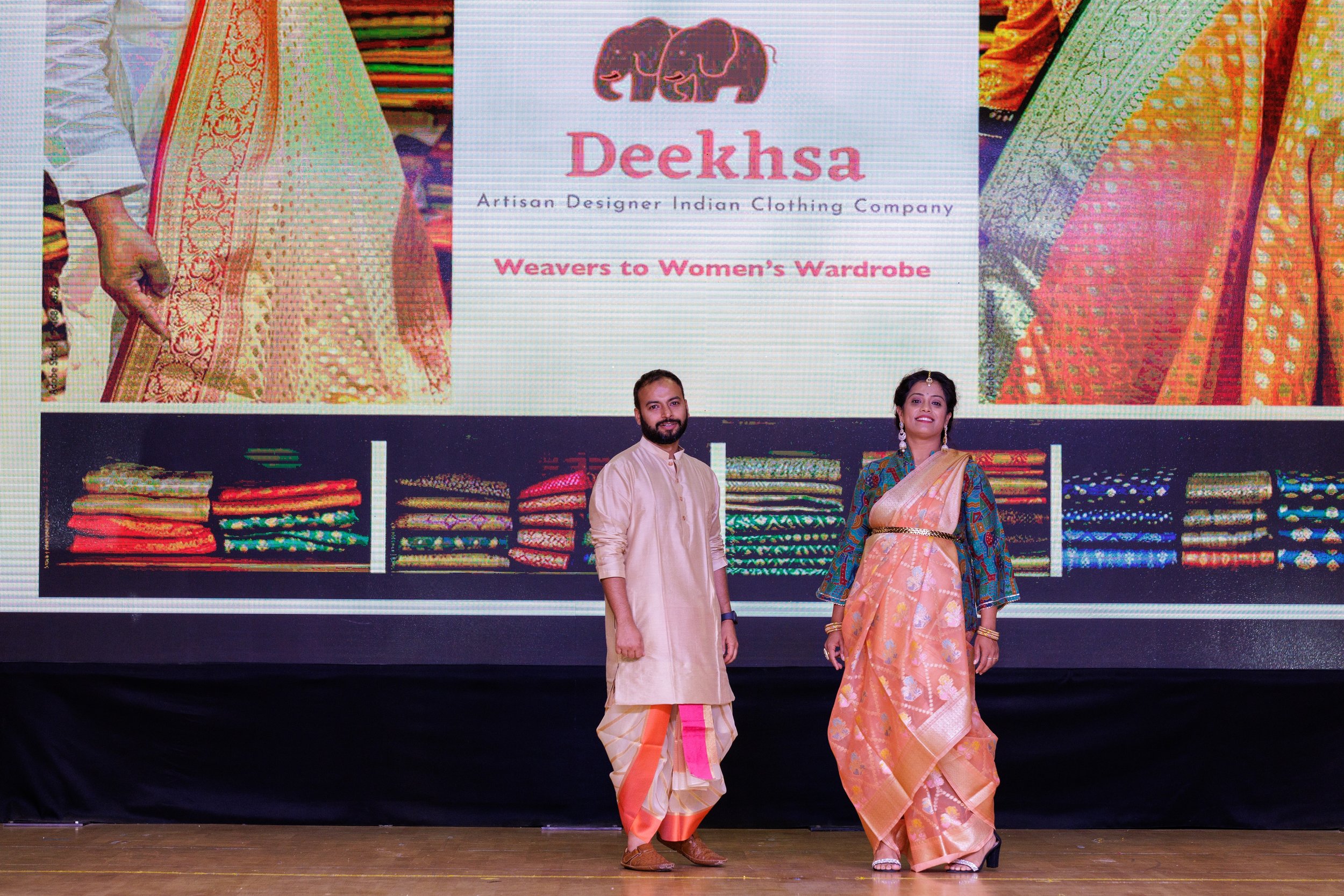 Two people standing on a stage in front of a large screen displaying colorful Indian textile designs and the logo for Deeksha, an artisan designer Indian clothing company. The man is wearing traditional cream-colored Indian attire with a peach and go