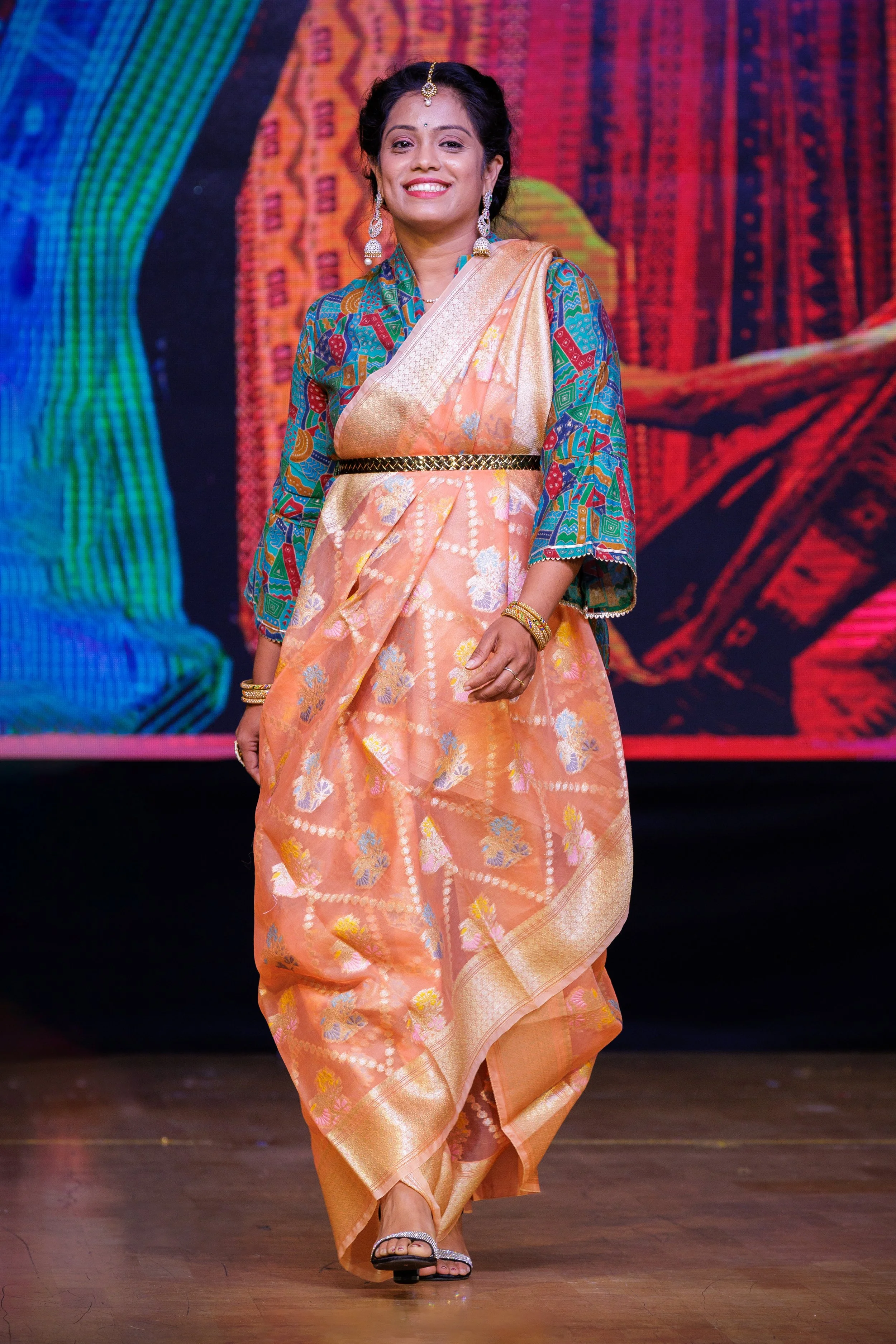 Woman in a colorful sari and earrings walking on stage with a vibrant fabric backdrop.