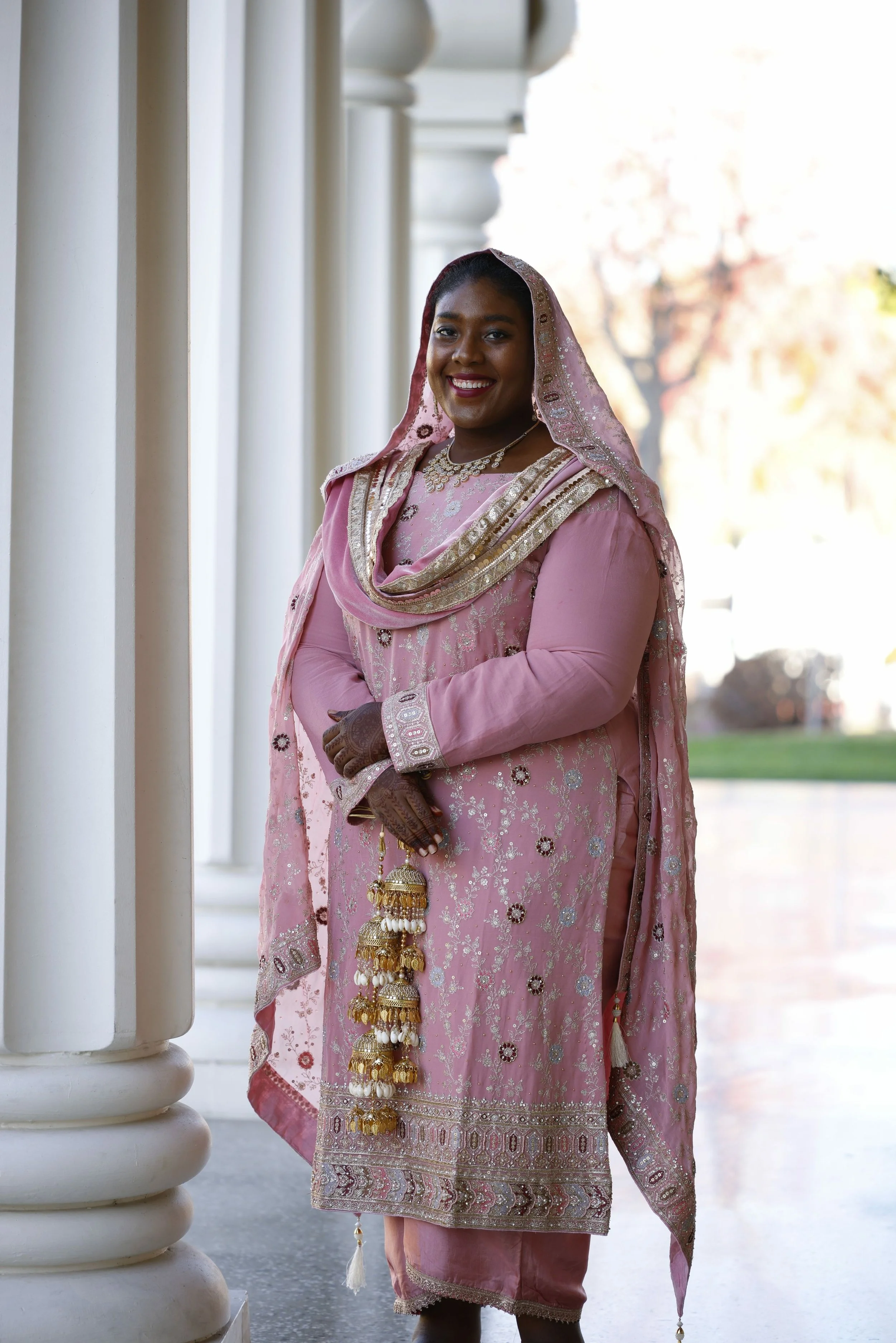 A woman dressed in traditional South Asian pink embroidered attire, standing outdoors by white columns, smiling, with a background of trees and sky.