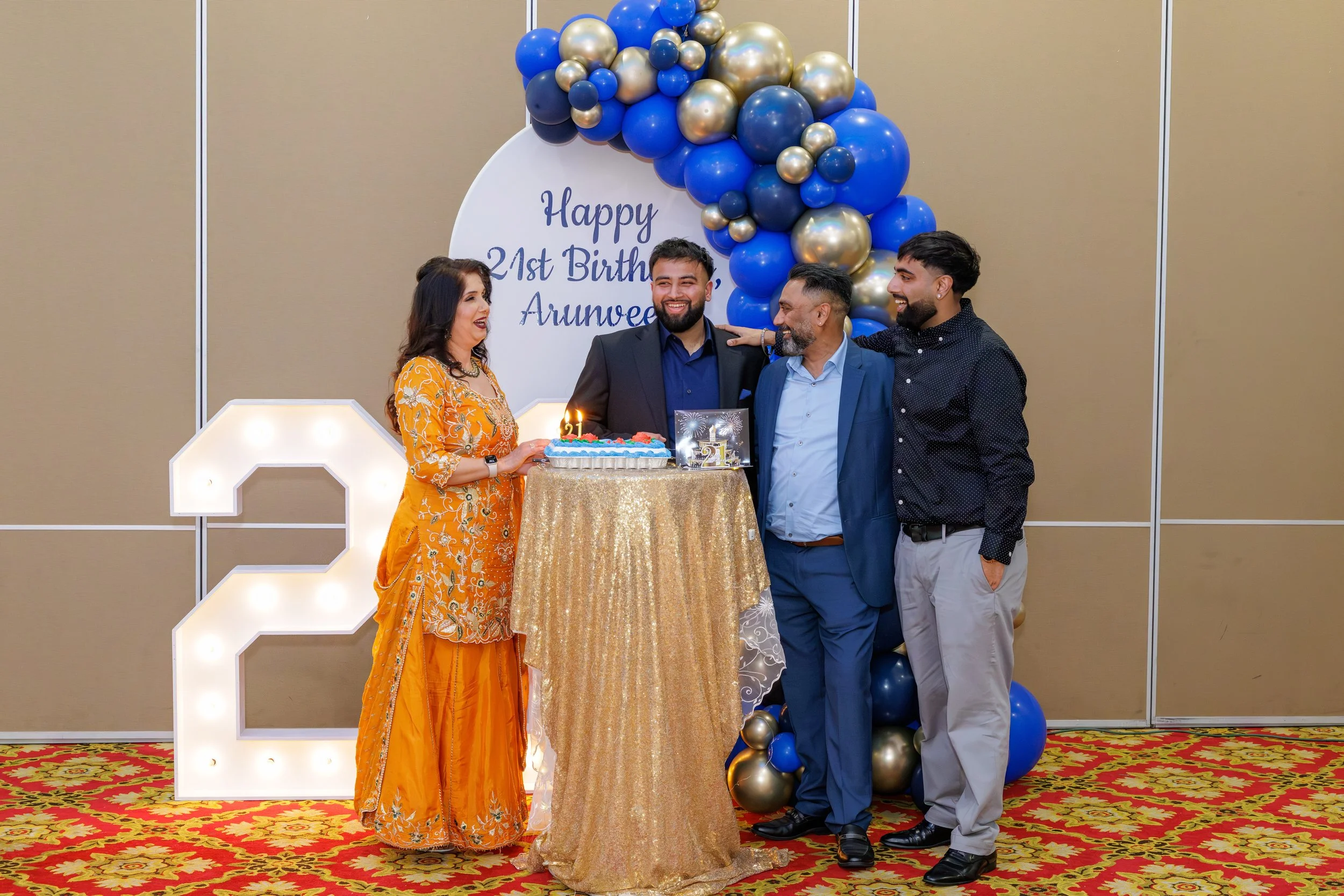 Group of four people celebrating a 21st birthday, standing around a cake on a gold tablecloth, with balloons and a large illuminated number 2 in the background, decorated with a sign reading 'Happy 21st Birthday Arunvee'.