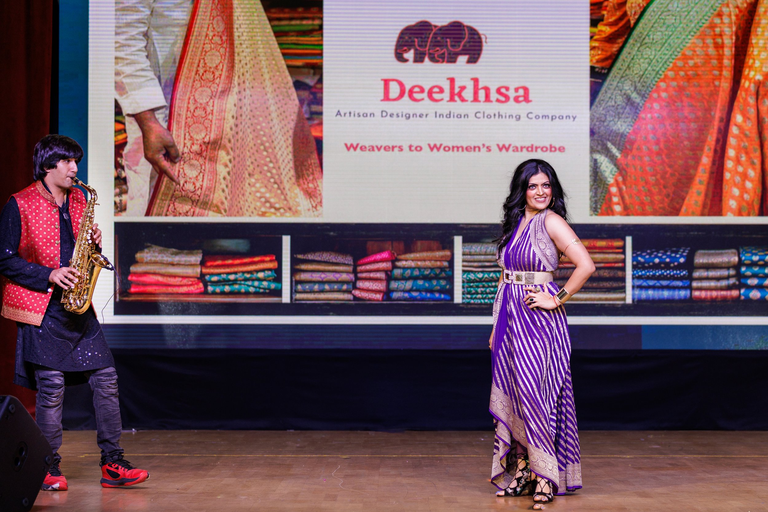 A woman in a purple and gold striped dress with a beige belt posing on stage in front of a large screen displaying a logo and images of Indian clothing. A musician playing the saxophone in red and black traditional attire is on her left.
