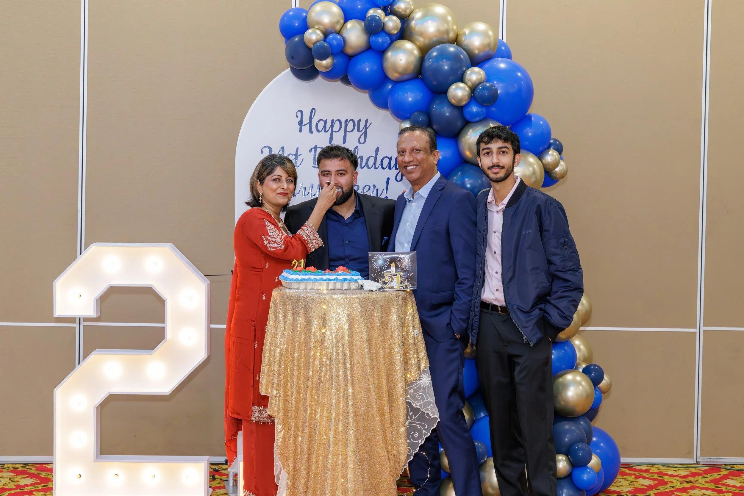 Four people celebrate a birthday with a cake, balloons, and a sign that reads 'Happy 21st Birthday.' They are standing in front of a decorated backdrop with blue, gold, and black balloons, and there is a large illuminated number two on the left side.