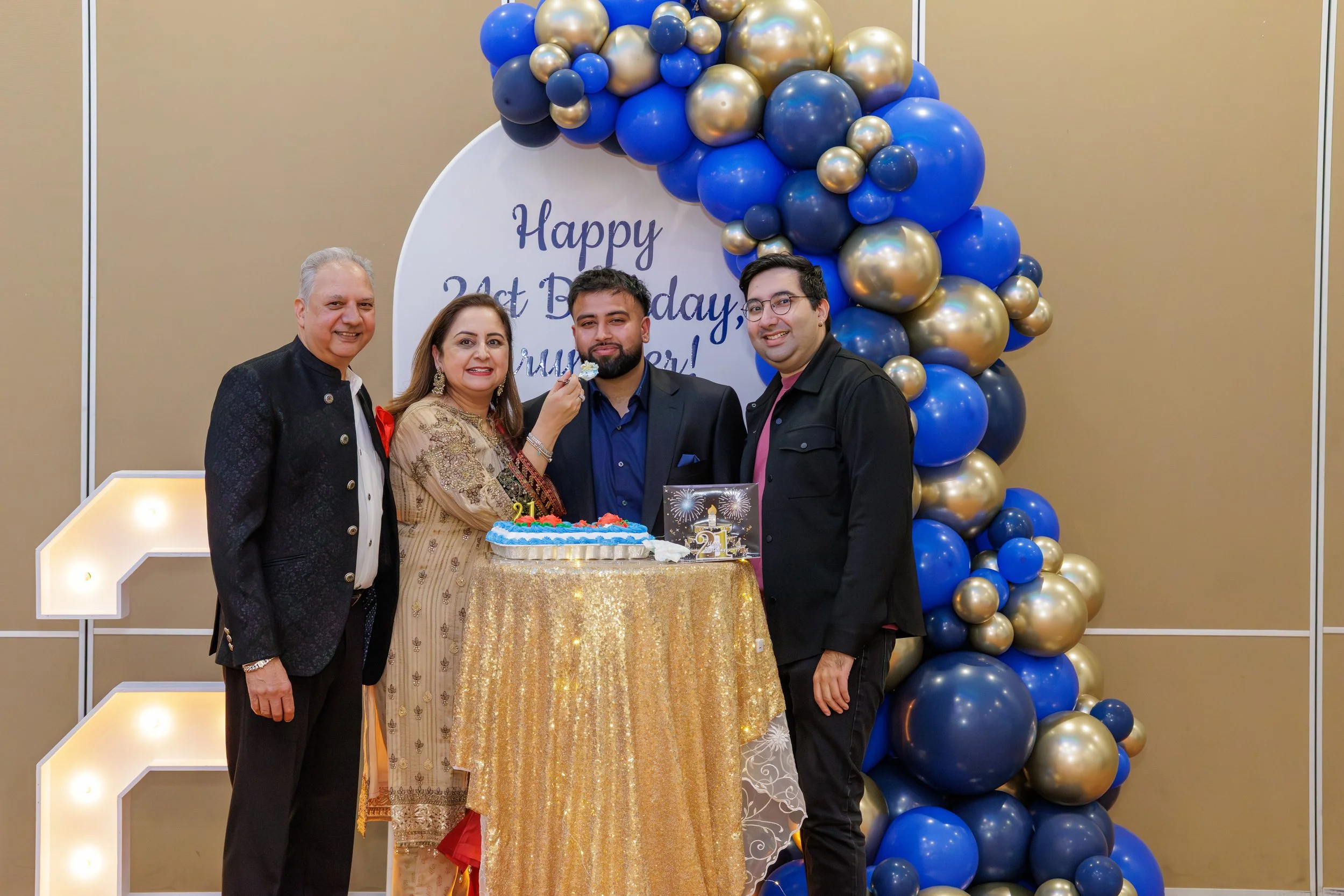 A family celebrating a 21st birthday, with four people standing around a table with a birthday cake, in front of a backdrop decorated with blue and gold balloons and a sign that reads 'Happy 21st Birthday, Mom!'