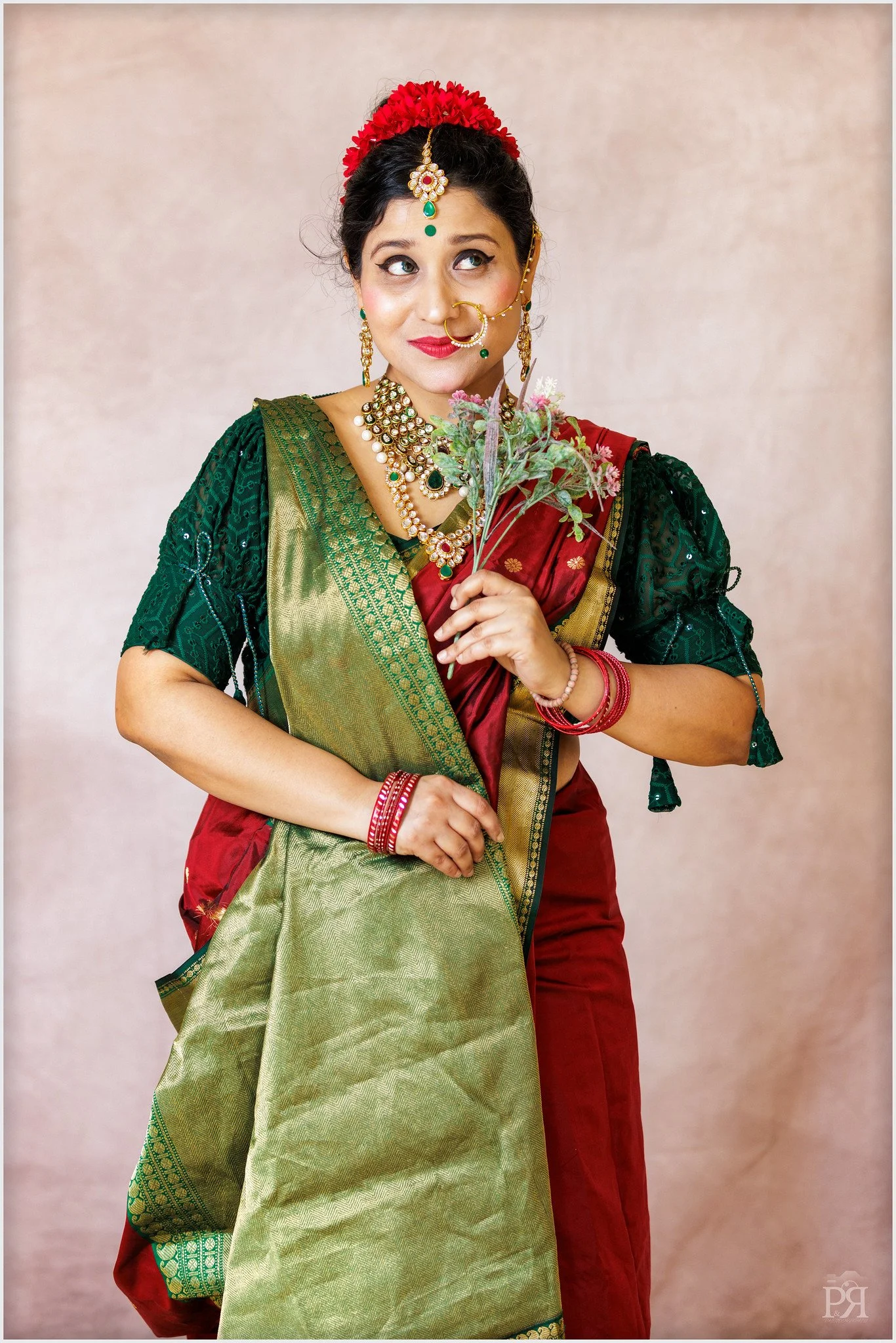 A woman dressed in traditional Indian attire, wearing a green blouse with intricate designs, a red saree with gold accents, and adorned with jewelry including necklaces, earrings, bangles, and a nose ring. She holds a small bouquet of pink flowers an