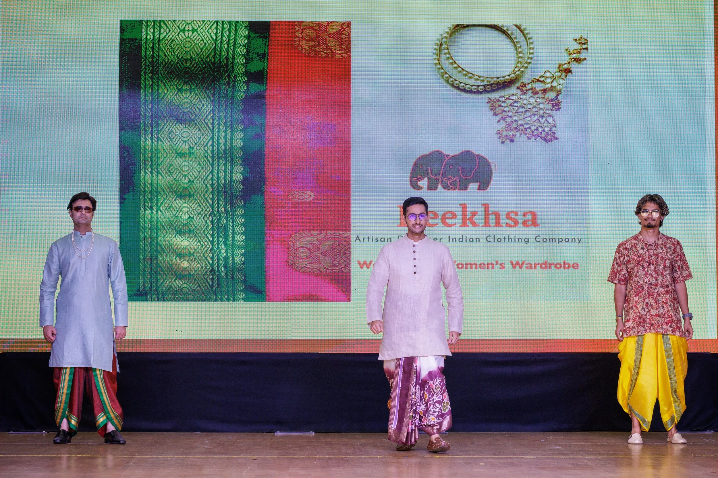 Three men in traditional Indian attire walking on a stage with a large screen behind them displaying an advertisement for Geekhsa, an artisan Indian clothing company. The screen features images of colorful fabric, jewelry, and the company logo.